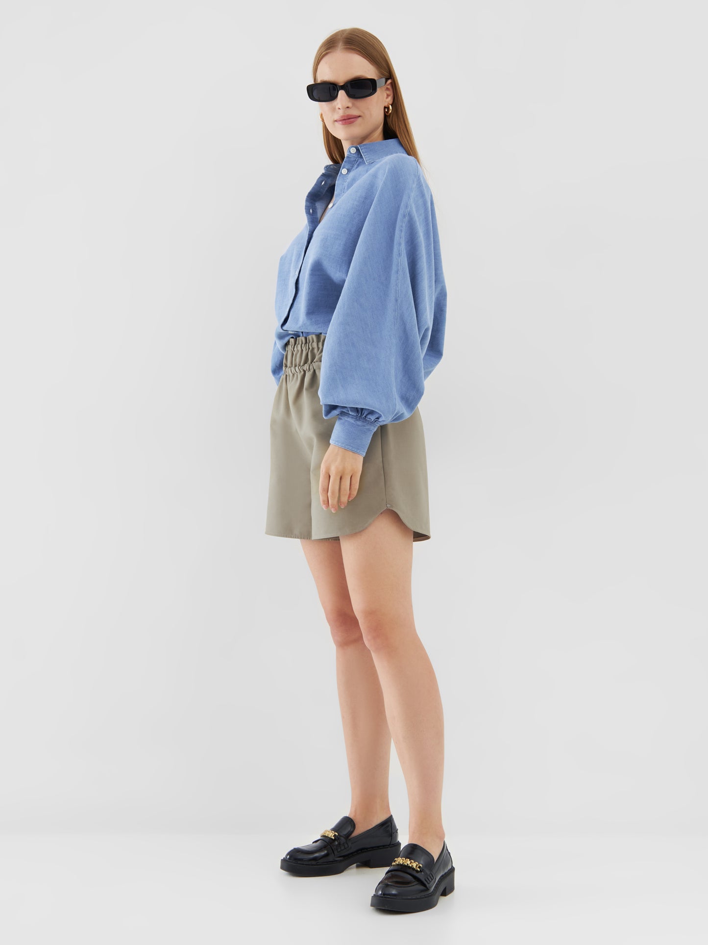 Eine Frau mit glattem Haar posiert selbstbewusst vor einem weißen Hintergrund und trägt eine hellblaue Bluse Antonia Slim Denim, beigefarbene Shorts mit hoher Taille, schwarze Slipper und eine Sonnenbrille.