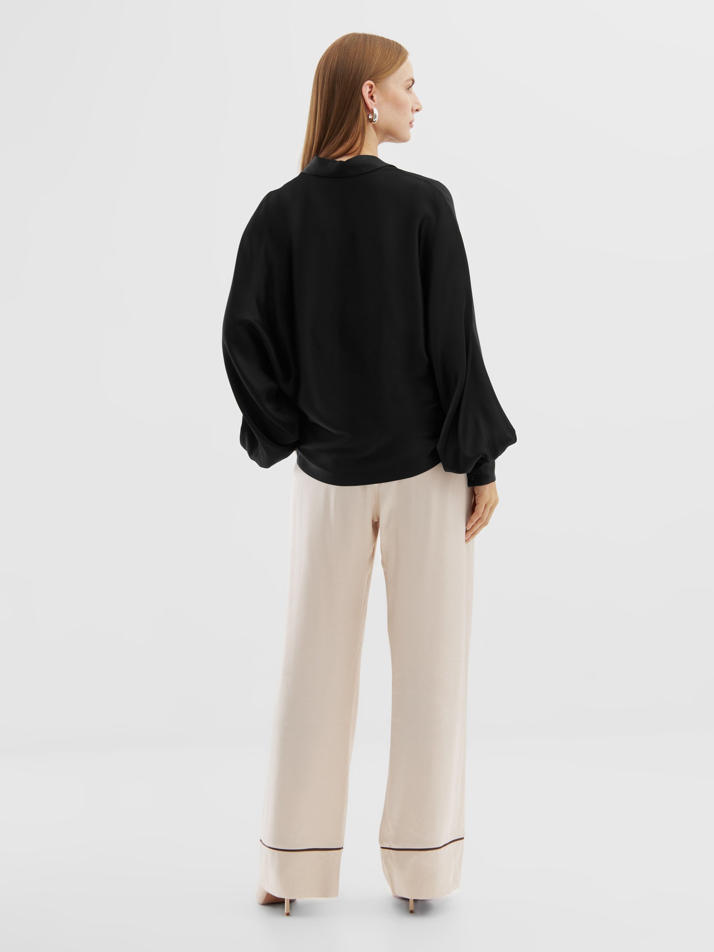 Eine Frau steht mit dem Gesicht zur Wand und trägt die Bluse Antonia Shiny Slim Black mit weiten Ärmeln und einer cremefarbenen Hose mit weitem Bein, die am Saum eingefasst ist. Sie hat glattes, hellbraunes Haar in einer minimalistischen Studioumgebung.