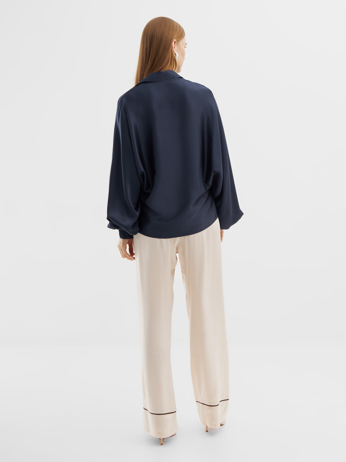 Eine Frau steht mit dem Gesicht zur Wand und trägt die Bluse Antonia Shiny Slim Navy mit langen Ärmeln und einer cremefarbenen Hose mit weitem Bein, die am Saum dunkel abgesetzt ist, vor einem schlichten weißen Hintergrund.