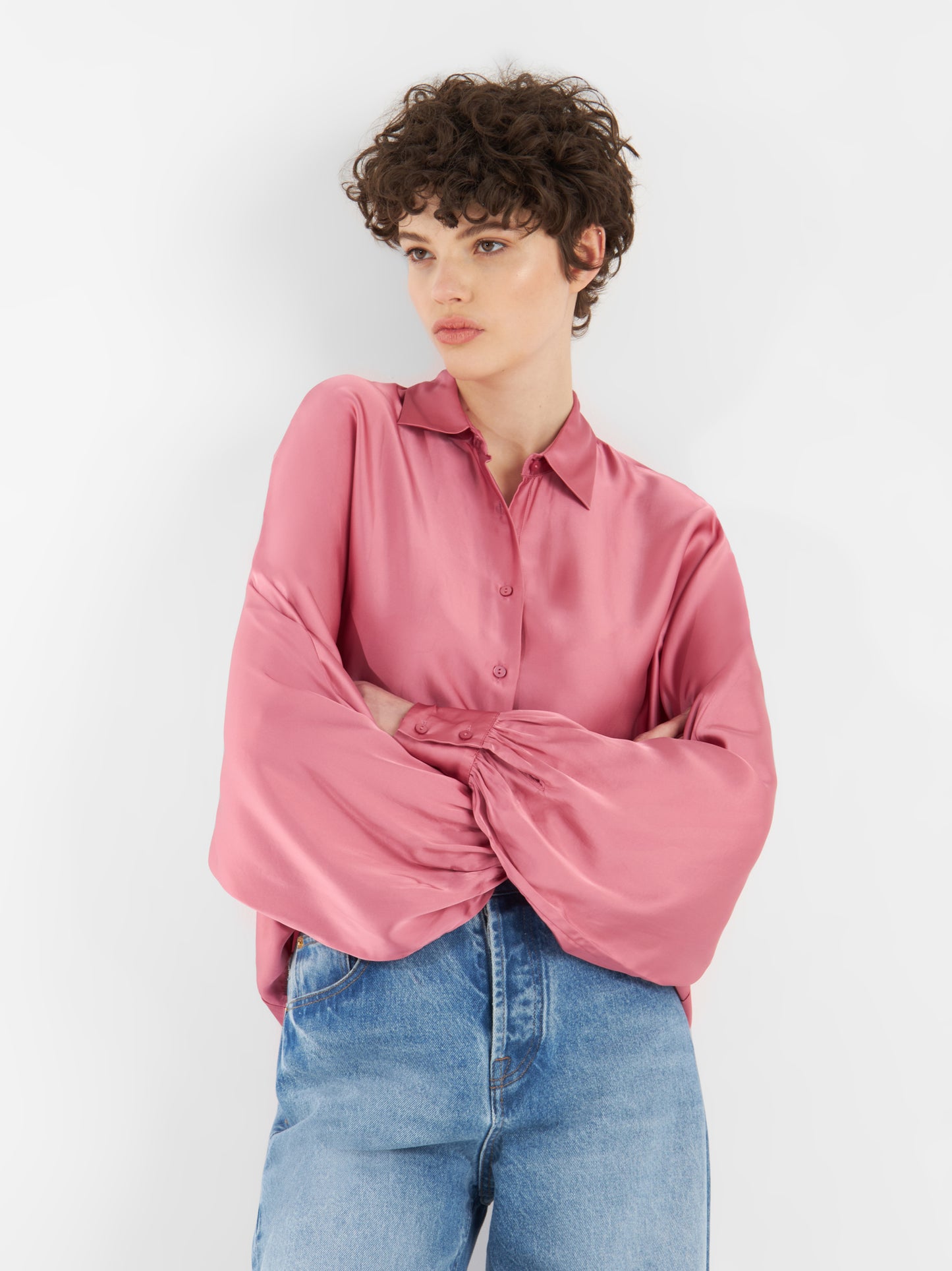 Eine Person mit kurzen, gelockten Haaren trägt eine Bluse Antonia Shiny Slim Rosewood mit weiten Ärmeln und Blue Jeans und steht mit verschränkten Armen vor einem schlichten weißen Hintergrund.