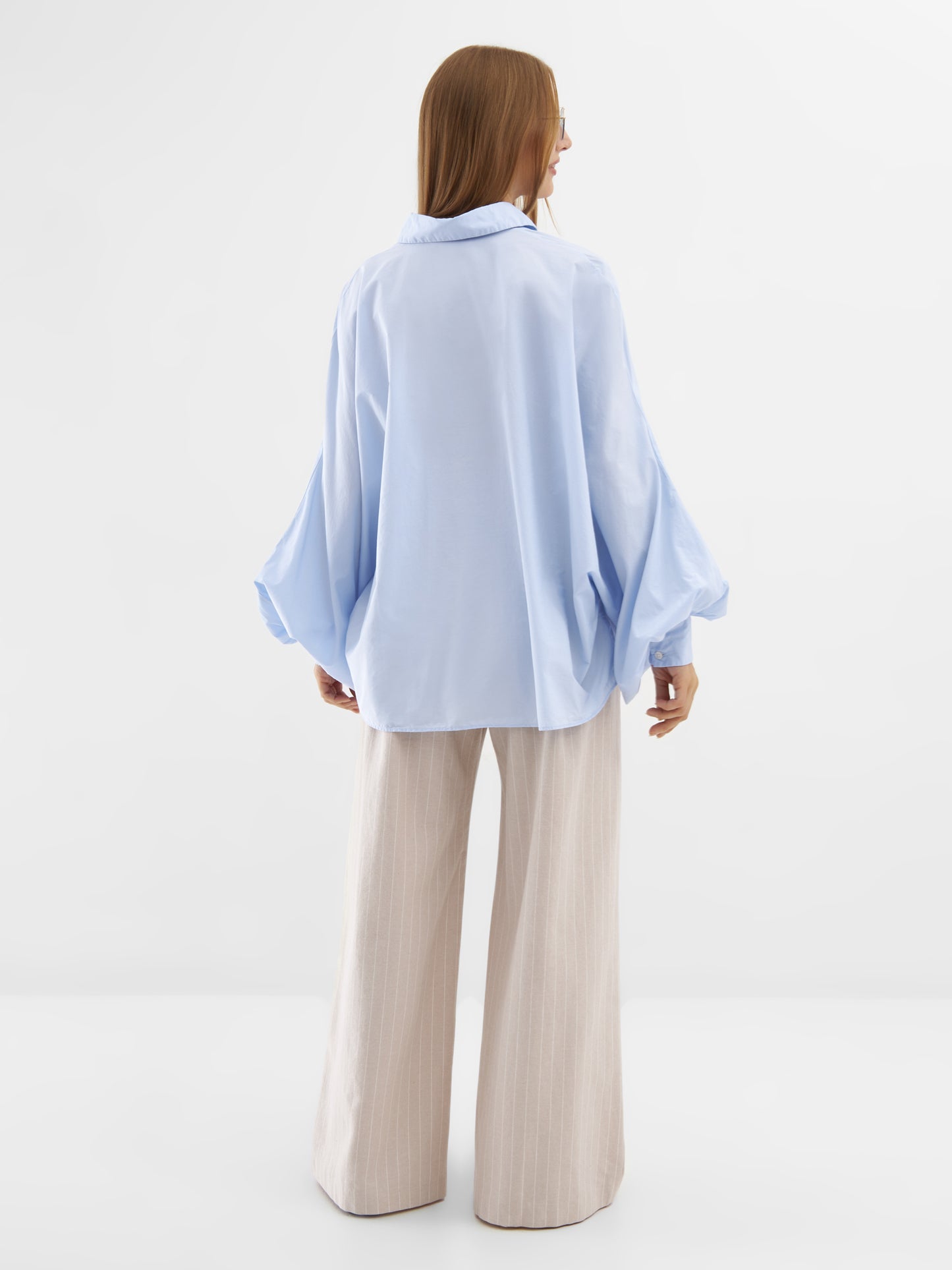 Eine Person mit langen hellbraunen Haaren steht mit dem Gesicht zur Wand und trägt die Bluse Antonia Lightblue in One Size mit weiten Ärmeln und beiger Hose mit weitem Bein vor einem schlichten weißen Hintergrund.