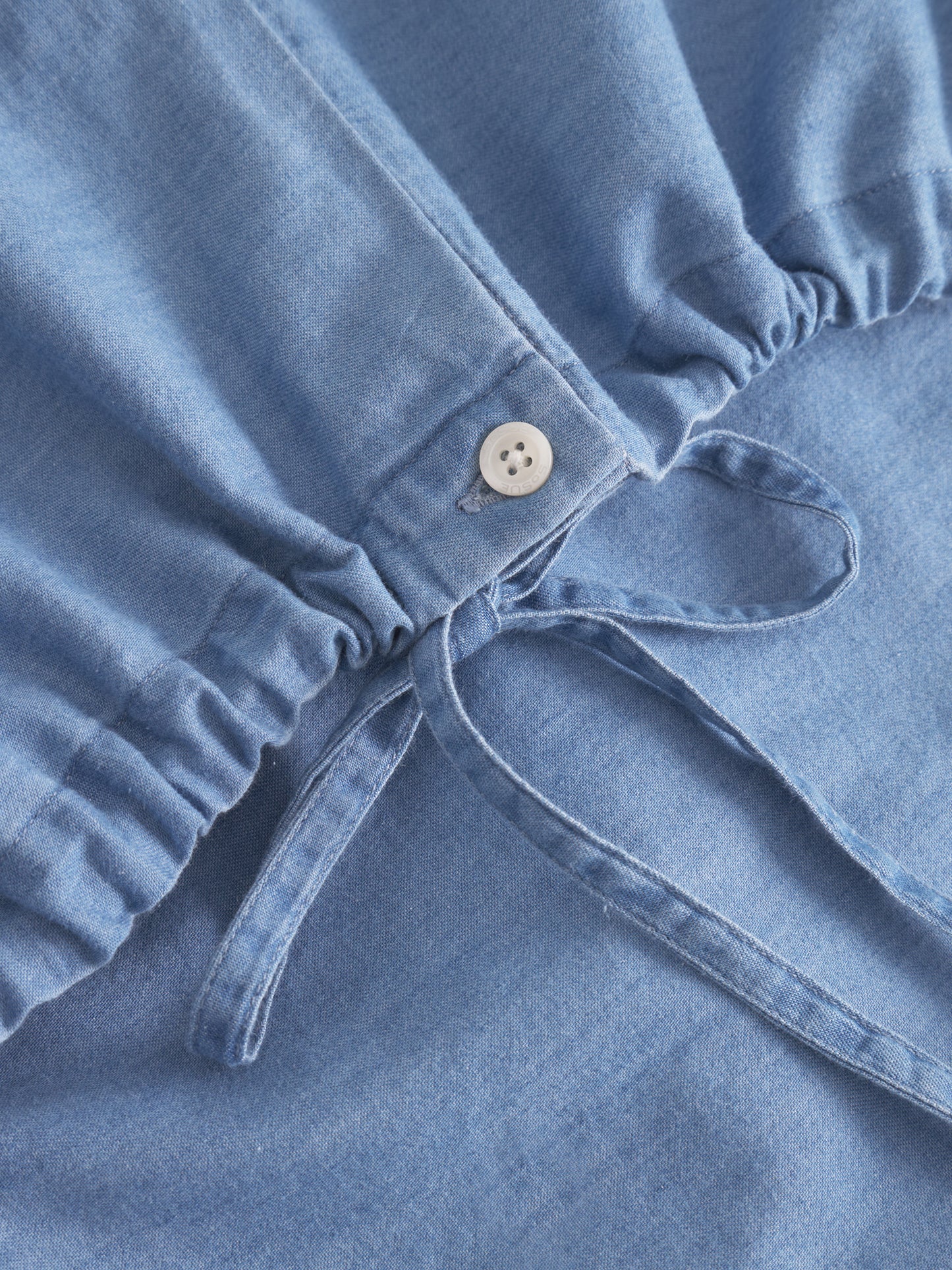 Nahaufnahme der Bluse Antonia Drawstring Denim in Hellblau, die einen Kordelzug aus Stoff und einen weißen Knopf an einer gerafften Naht zeigt. Die weiche Denim-Textur sticht hervor und unterstreicht den Oversized-Look für Damen.