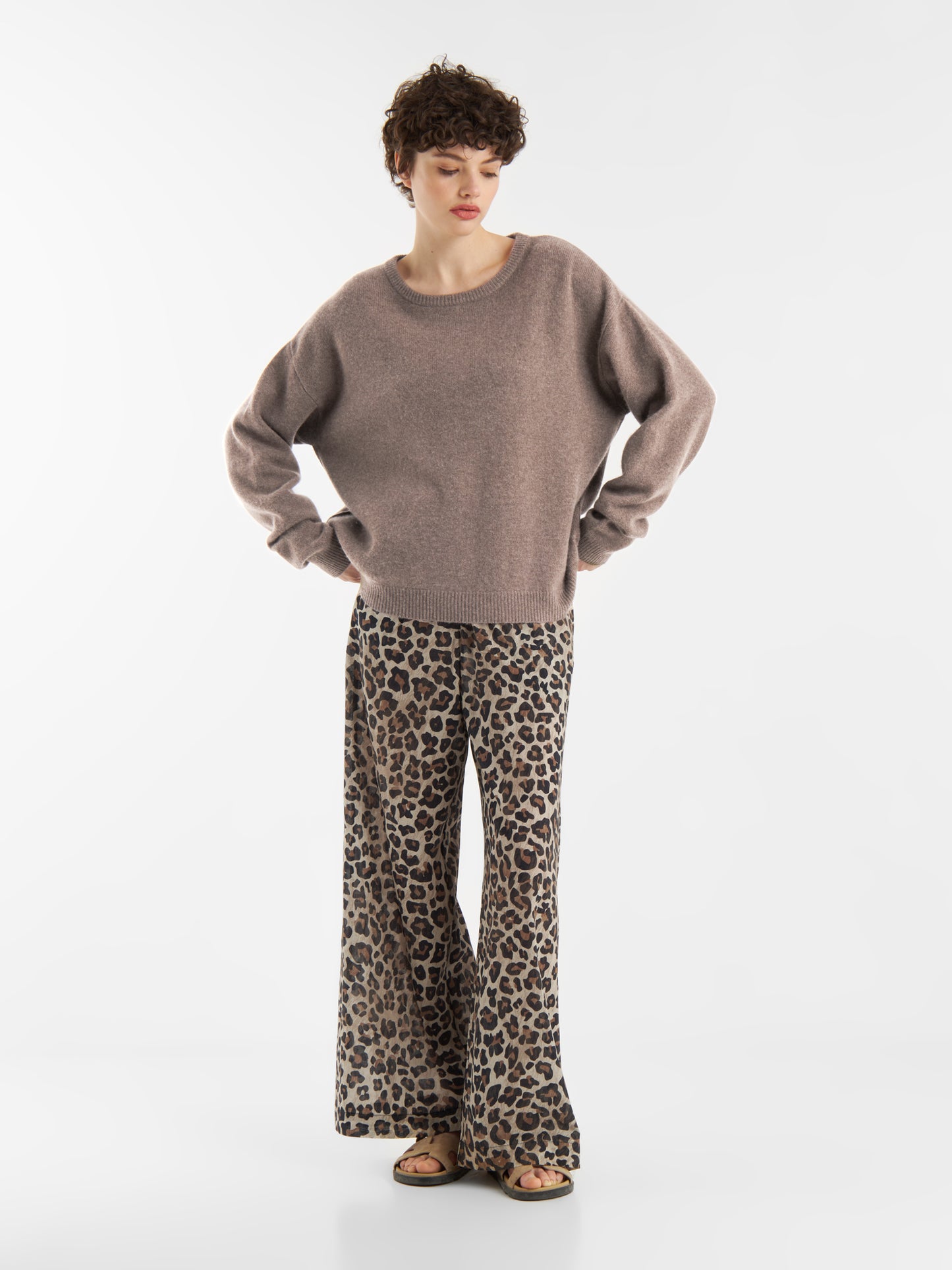 Person mit kurzen lockigen Haaren steht vor einem unifarbenen Hintergrund und trägt einen lockeren Pullover Boyfriend azteco mit Rundhalsausschnitt, eine Hose mit weitem Bein im Leopardenmuster und Sandalen, die Hände in die Hüften gestemmt.