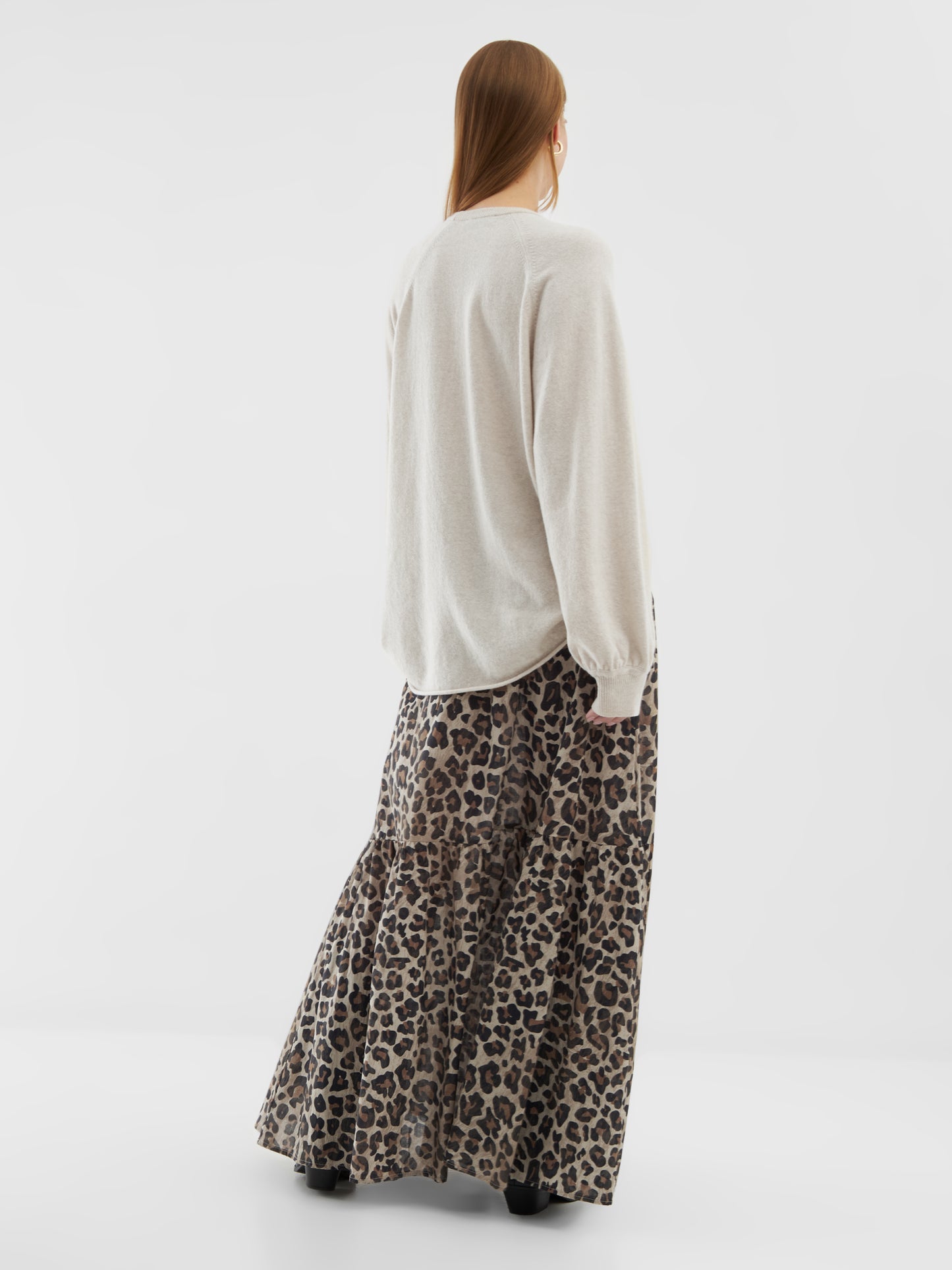 Eine Person mit langen Haaren blickt in die Ferne. Sie trägt den Pullover Elli Beige und einen langen, gestuften Rock mit Leopardenmuster und schwarzen Stiefeln vor einem schlichten weißen Hintergrund.