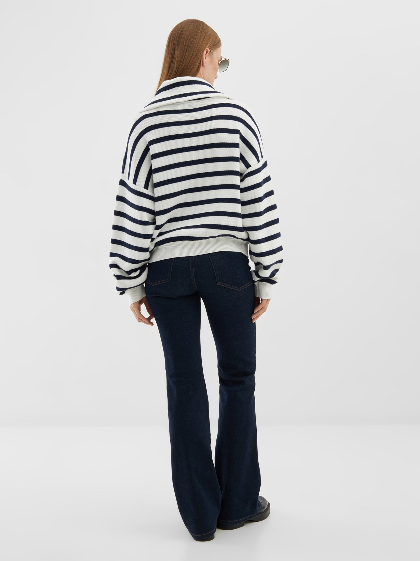 Eine Person mit langem, glattem Haar, die eine Sonnenbrille, einen Pullover mit Reißverschluss, dunkelblaue Schlagjeans und schwarze Schuhe trägt, steht mit dem Gesicht nach unten vor einem schlichten weißen Hintergrund.