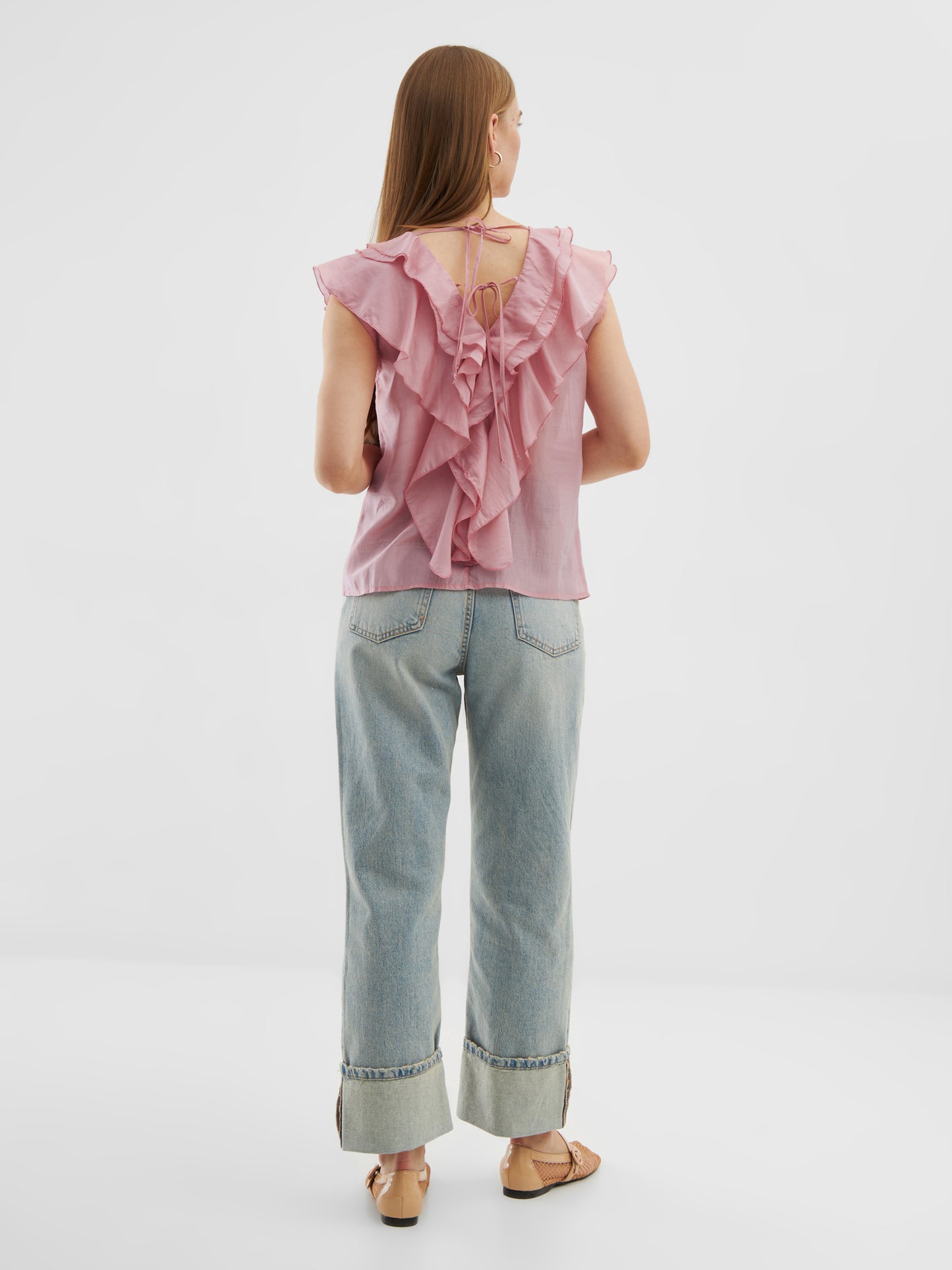 Eine Frau, von hinten gesehen, trägt das Top Ruffle Light Pink mit V-Ausschnitt, hellblaue Jeans mit Bündchen und Sandalen. Ihr langes Haar fällt vor einem schlichten weißen Hintergrund nach unten.