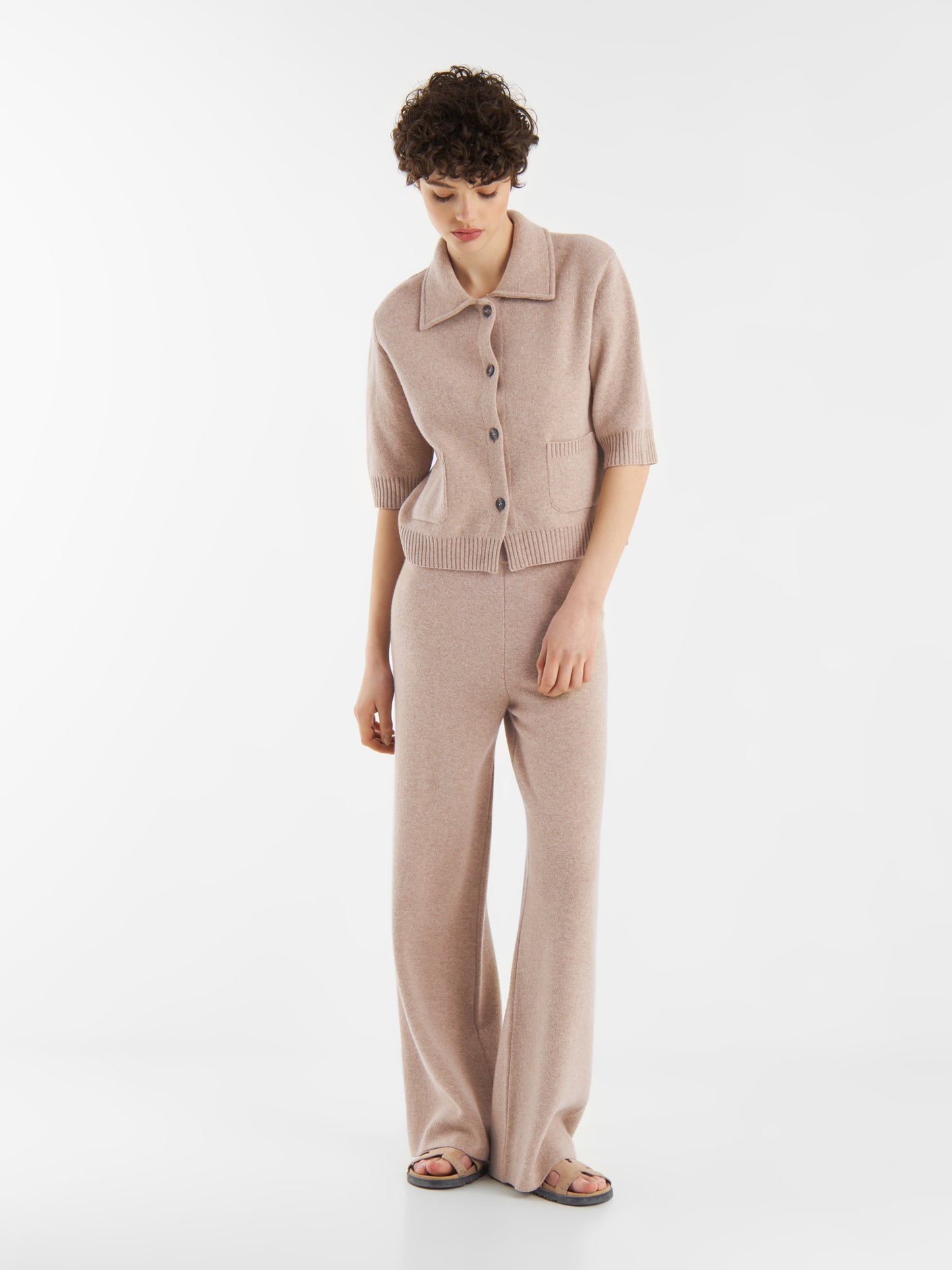 Eine Person mit kurzen lockigen Haaren trägt ein hellrosafarbenes, kurzärmeliges, geknöpftes Strickoberteil und eine dazu passende Hose Merino Beige mit elastischem Bund und weiten Beinen, die vor einem schlichten weißen Hintergrund steht.