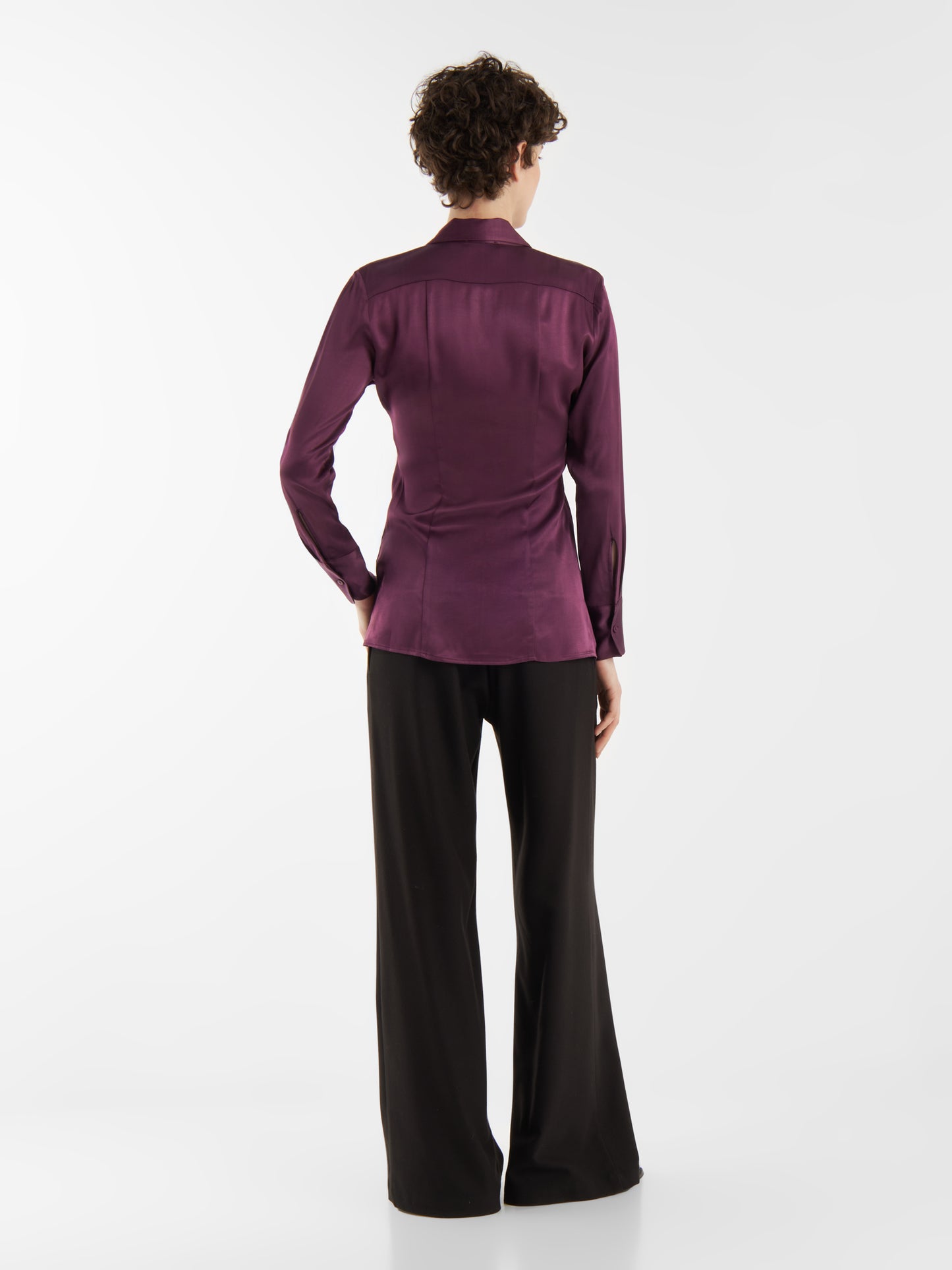 Eine Person mit kurzen lockigen Haaren steht mit dem Gesicht zur Wand und trägt die Bluse Ella Shiny Purple und eine schwarze Hose mit weitem Bein vor einem schlichten weißen Hintergrund.