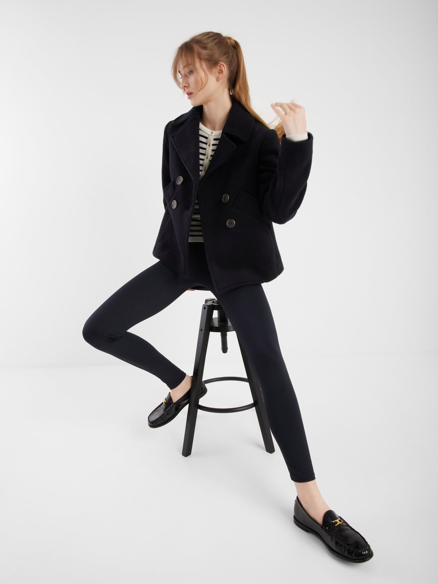 Eine Frau sitzt auf einem hohen schwarzen Hocker und trägt die Jacke Caban Navy, schwarze Leggings, ein gestreiftes Top und schwarze Halbschuhe. Sie blickt mit einem angewinkelten Knie zur Seite und hält eine Haarsträhne vor einen schlichten weißen Hintergrund.