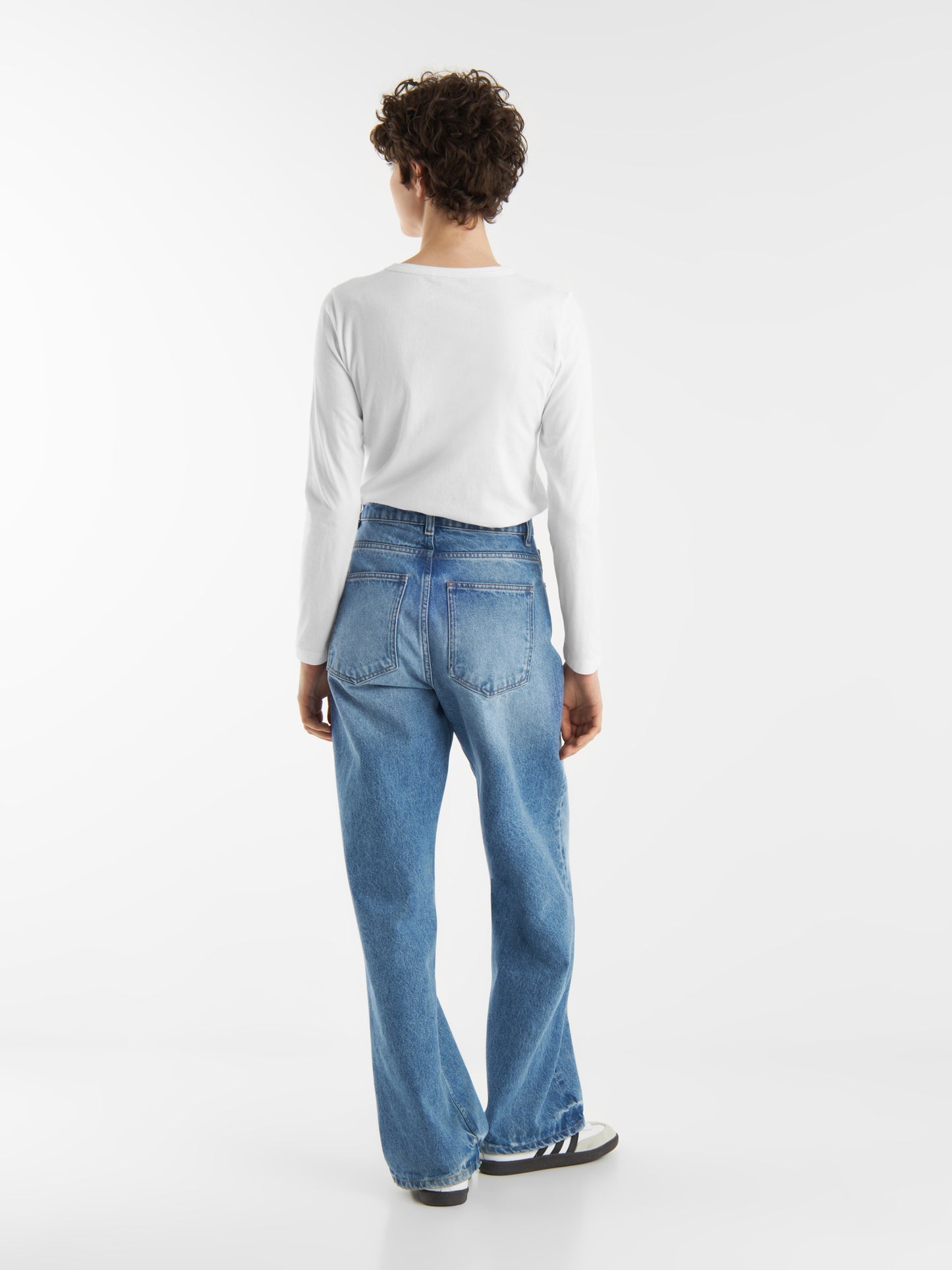 Eine Person mit kurzen lockigen Haaren, die ein langärmeliges weißes Hemd, blaue Jeans mit hoher Taille und schwarz-weiße Turnschuhe trägt, steht mit dem Gesicht nach hinten vor einem schlichten weißen Hintergrund.