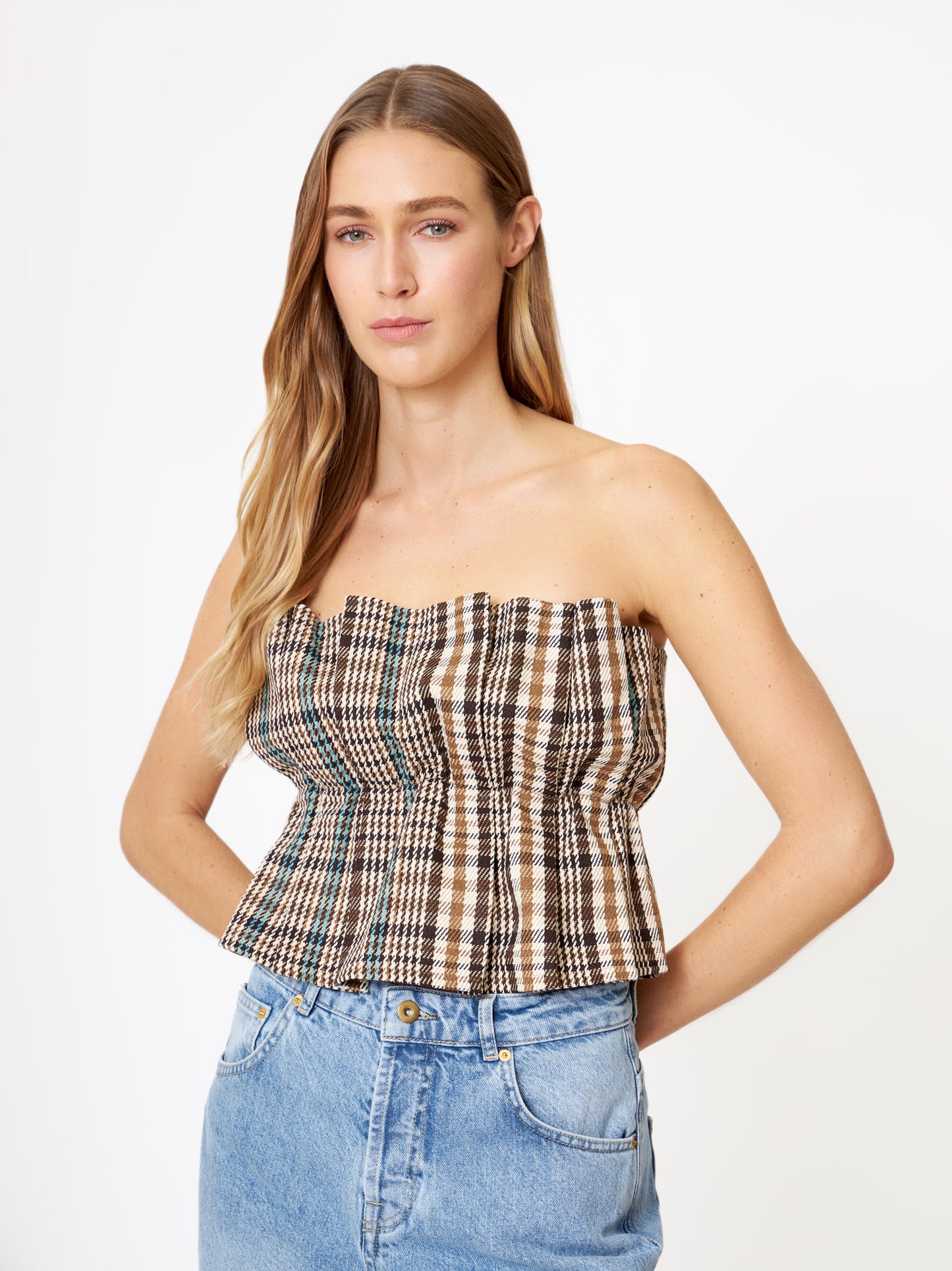 Eine Frau mit langem, hellbraunem Haar trägt das Pleat Top Check und blaue Jeans und steht mit neutralem Gesichtsausdruck vor einem schlichten weißen Hintergrund.