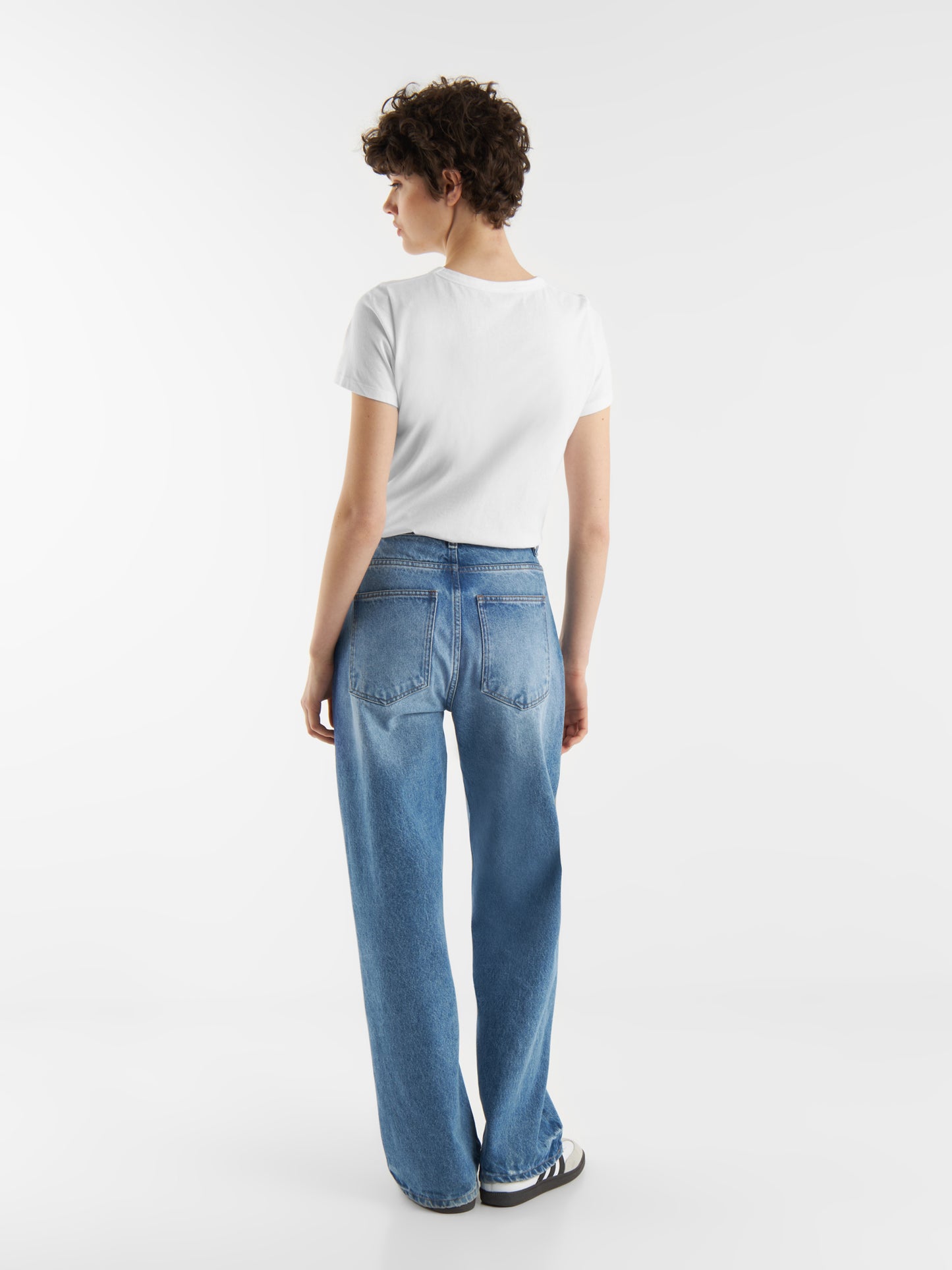 Eine Person mit kurzen lockigen Haaren trägt das T-Shirt Roundneck White, weite Bluejeans und schwarz-weiße Turnschuhe und steht mit dem Rücken zu einem schlichten weißen Hintergrund.