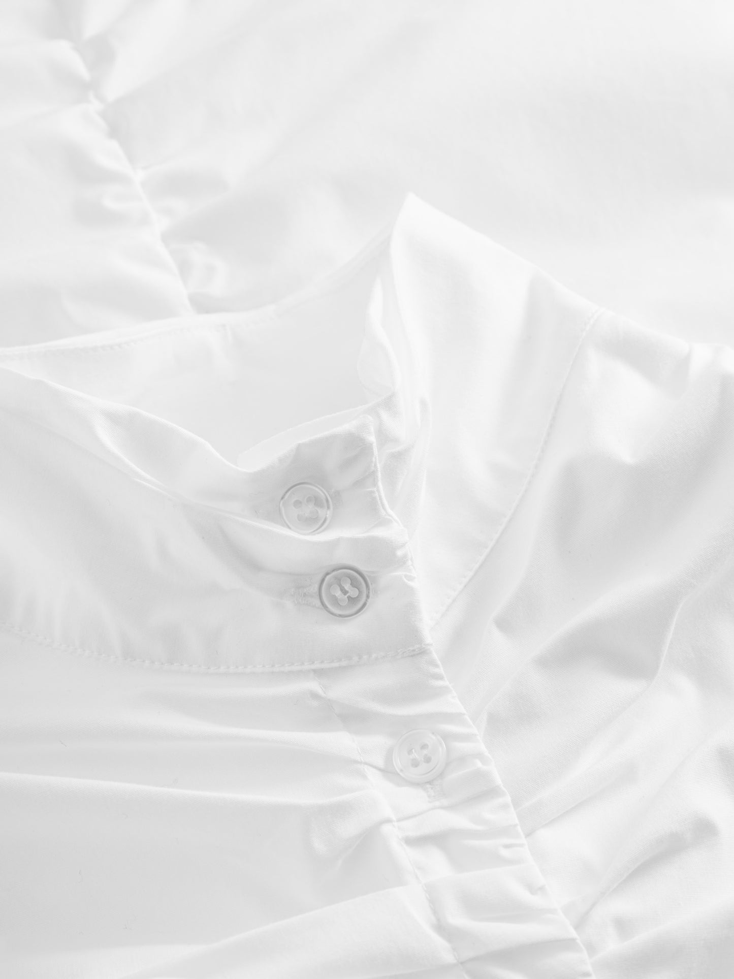 Nahaufnahme der Bluse Virginy white, eine elegante Damenbluse mit Stehkragen und drei weißen Knöpfen, leicht gekräuseltem Stoff und sanfter Beleuchtung, die die glatte Textur betont.