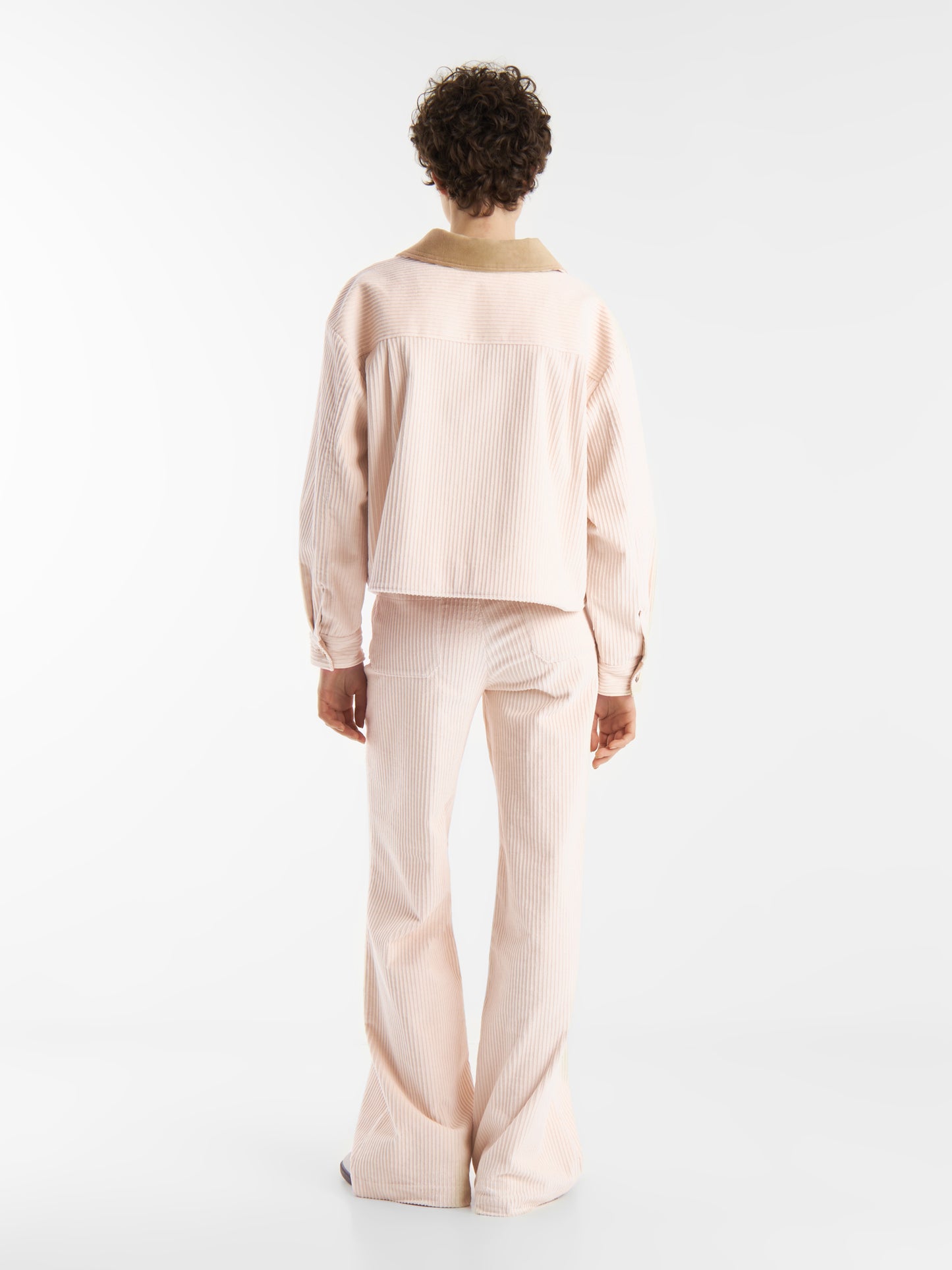 Person mit kurzen lockigen Haaren trägt eine hellrosa Cordjacke OFW und eine passende Schlaghose, steht vor einem schlichten weißen Hintergrund und blickt von der Kamera weg.