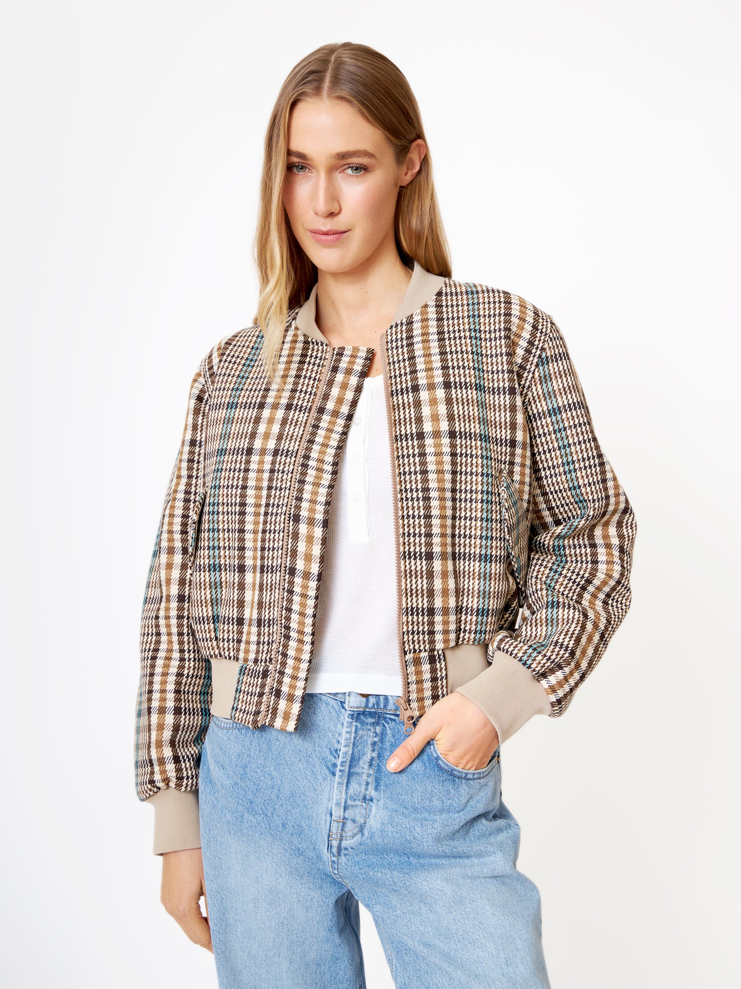 Eine Frau mit langen blonden Haaren trägt die Jacke Bomber Check über einem weißen Oberteil und hellblauen Jeans. Sie posiert vor einem schlichten weißen Hintergrund, hat eine Hand in der Tasche und schaut leicht lächelnd in die Kamera.