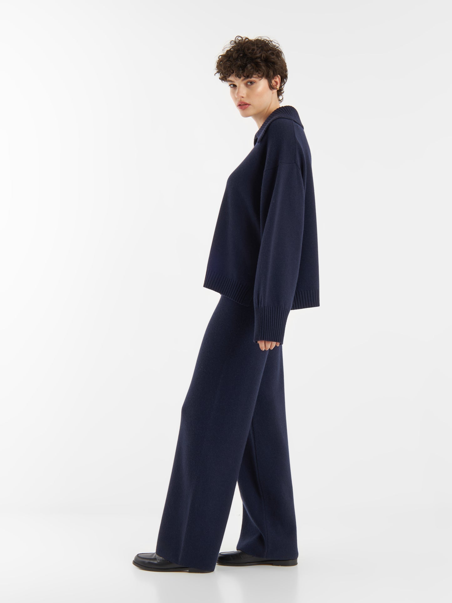 Eine Person mit kurzen lockigen Haaren, bekleidet mit einem lockeren dunkelnavyfarbenen Pullover und einer dazu passenden Hose Merino Darklblue, steht seitlich vor einem schlichten weißen Hintergrund.