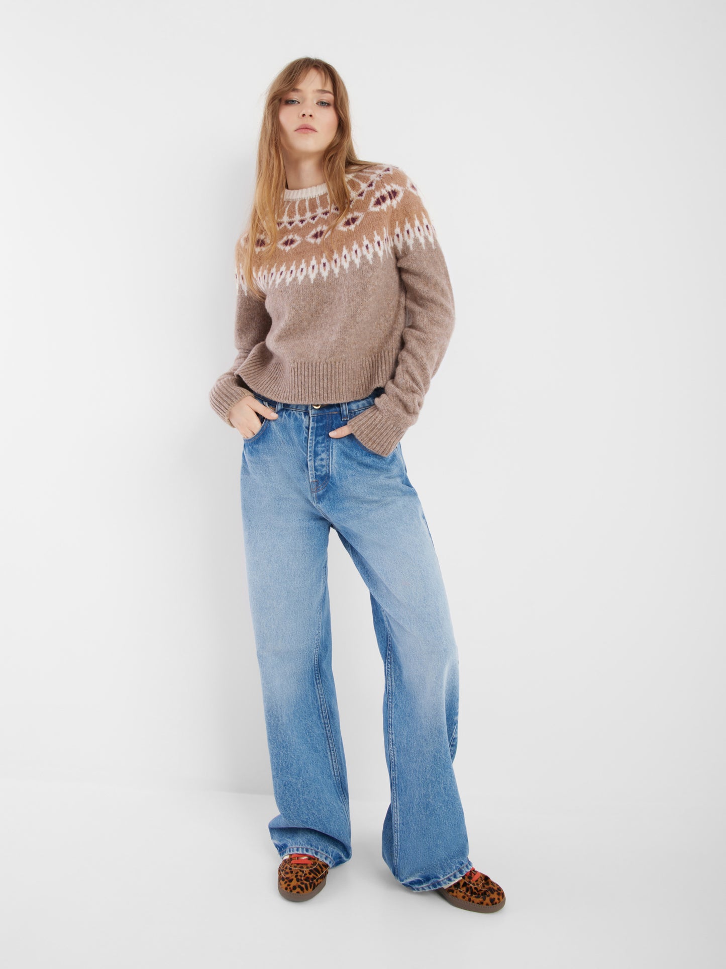 Eine Frau mit langen, glatten Haaren posiert vor einem weißen Hintergrund und trägt den Pullover Norweger Vol.2 Beige Burgundy, weite Bluejeans und flache Schuhe mit Leopardenmuster. Ihre Hände stecken in ihren Taschen.