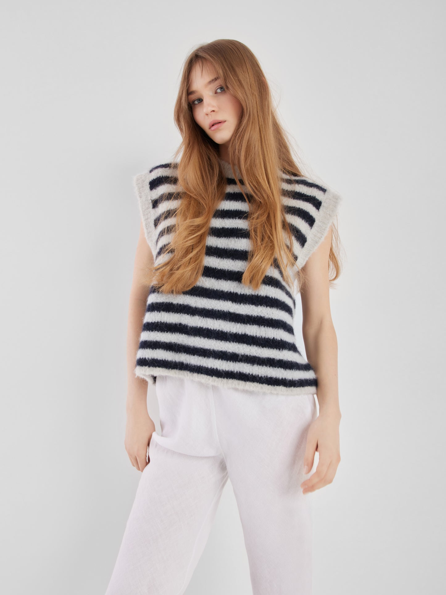 Eine Frau mit langem, hellbraunem Haar trägt einen Pullunder Blue Stripe und eine weite weiße Hose und steht vor einem schlichten hellgrauen Hintergrund.