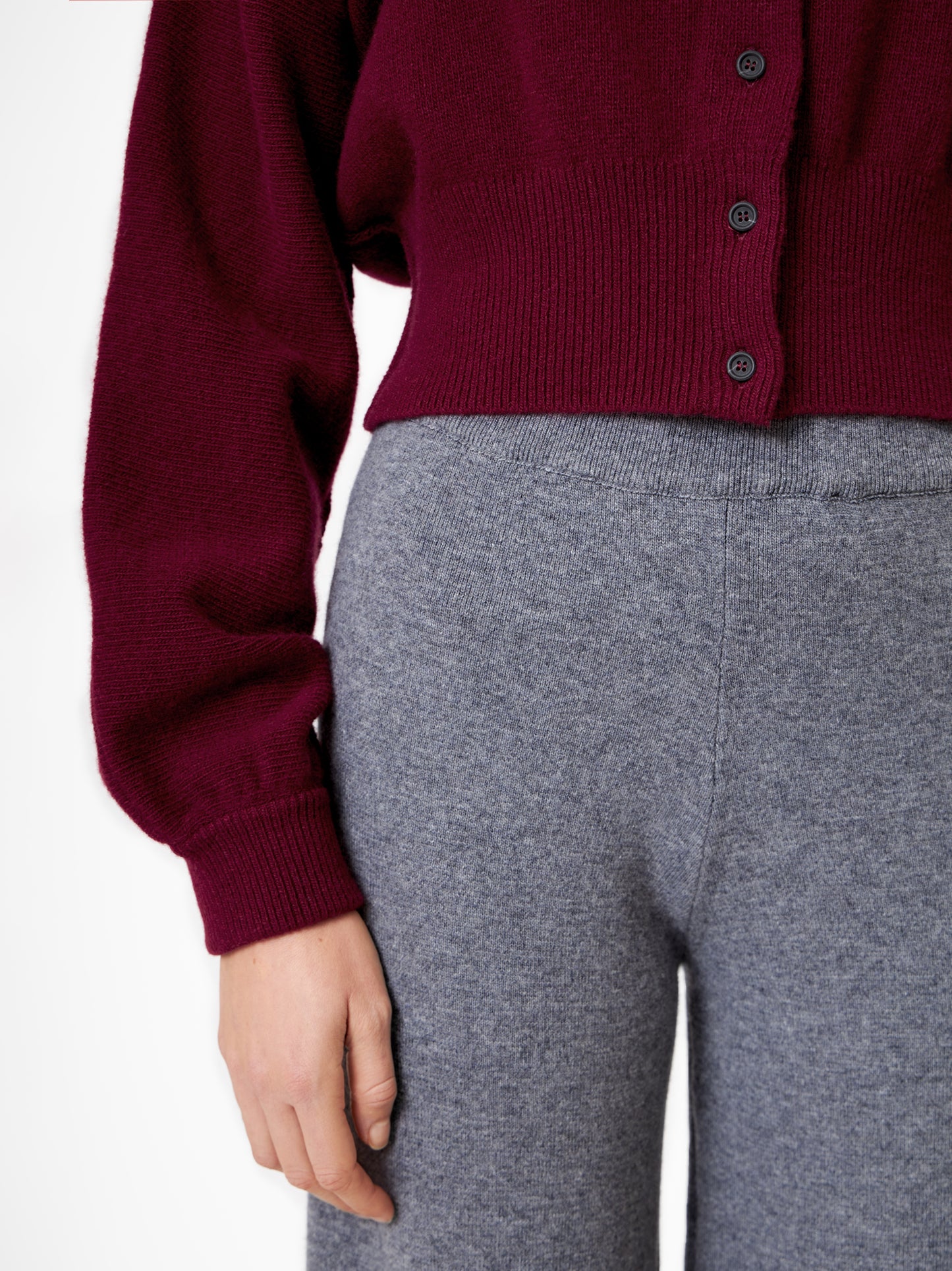 Vor einem unifarbenen Hintergrund ist eine Person von den Schultern bis zur Mitte des Oberschenkels abgebildet, die eine Hose in Merinograu und einen bordeauxfarbenen Pullover mit Knöpfen trägt, wobei die linke Hand entspannt an der Seite liegt.