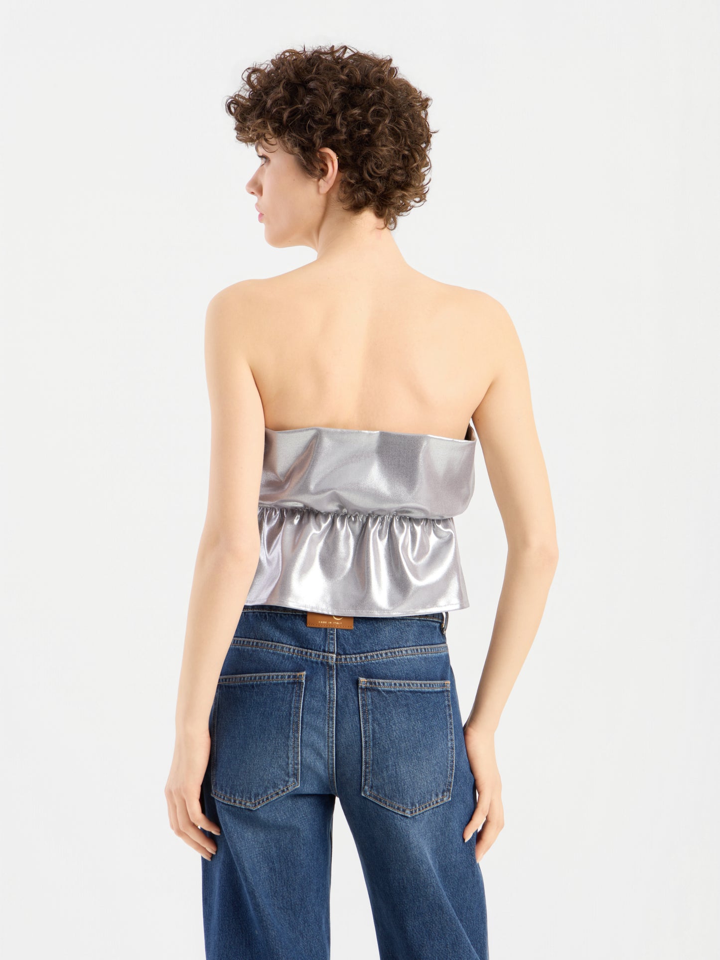 Eine Person mit kurzen Locken trägt ein „Pleat Top Silber“ und eine blaue Jeans und steht mit dem Rücken zur Kamera vor einem schlichten weißen Hintergrund.