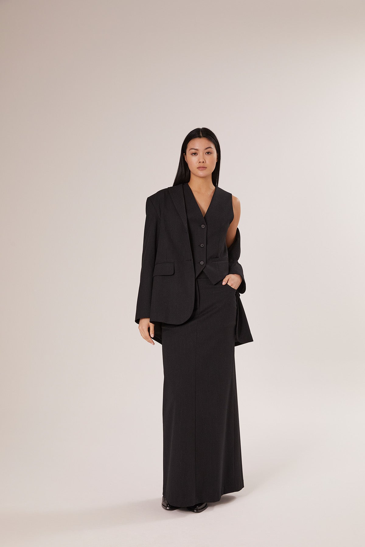 Eine Frau mit langen dunklen Haaren steht vor einem schlichten Hintergrund und trägt den Blazer Pinstripe Black als Teil eines schicken dreiteiligen Anzugs, gepaart mit einer ärmellosen Weste und einem langen Rock, selbstbewusst in die Kamera.
