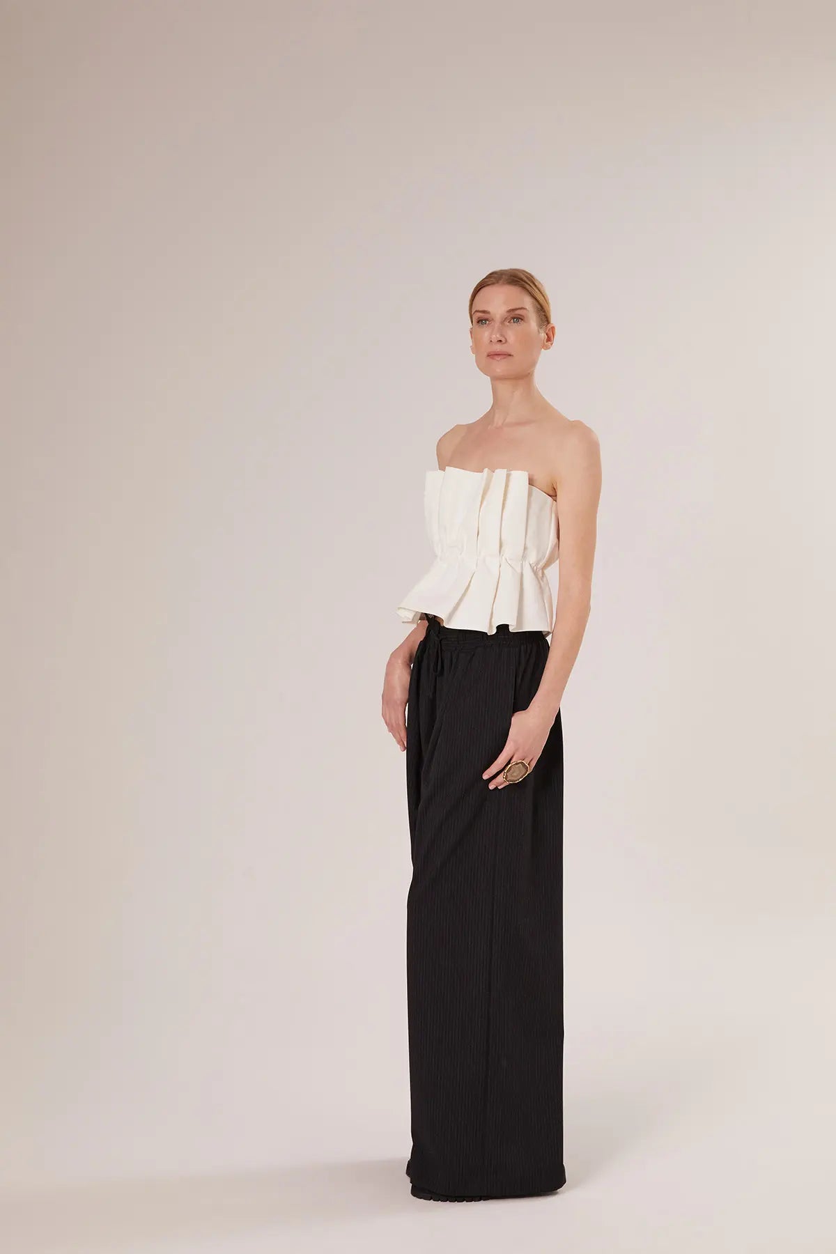 Eine Frau steht vor einem schlichten Hintergrund und trägt das Pleat Top White - ein schulterfreies weißes Crop-Top mit plissierten Details - zu einer schwarzen Hose mit hoher Taille und weitem Bein. Ihr Haar ist zurückgesteckt und sie hat einen neutralen Gesichtsausdruck.