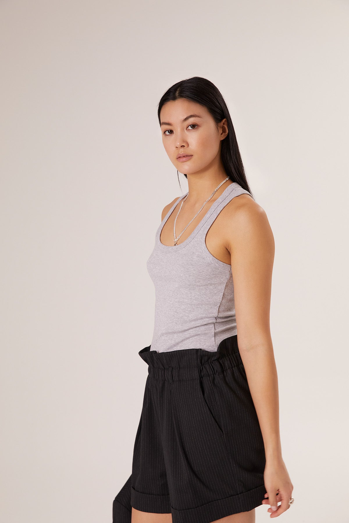 Eine Frau mit langen schwarzen Haaren trägt das Tank-Top „Rib Grey“ aus Baumwolle für Damen und schwarze, hoch taillierte Shorts – ein stilvolles Beispiel für nachhaltige Damenmode – steht vor einem schlichten, hellen Hintergrund und blickt in die Kamera.