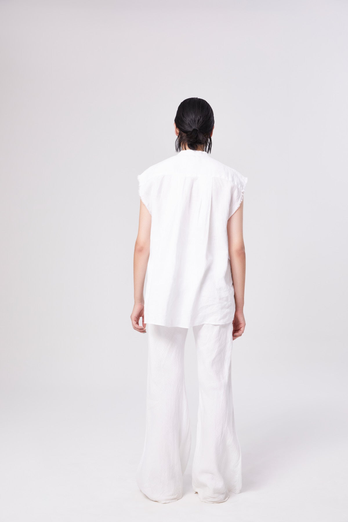 Eine Person mit dunklem, schulterlangem Haar steht mit dem Gesicht nach unten, trägt ein ärmelloses weißes Oberteil und die Hose Joana White vor einem schlichten weißen Hintergrund.