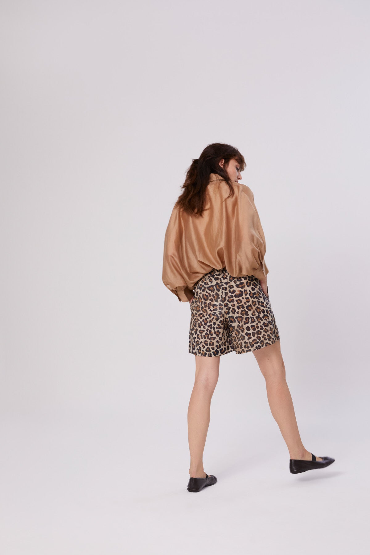 Eine Person mit mittellangen braunen Haaren steht abgewandt und trägt die Bluse Antonia Short Sleeve Gold sowie Shorts mit Leopardenmuster und schwarzen Flats vor einem schlichten hellen Hintergrund.