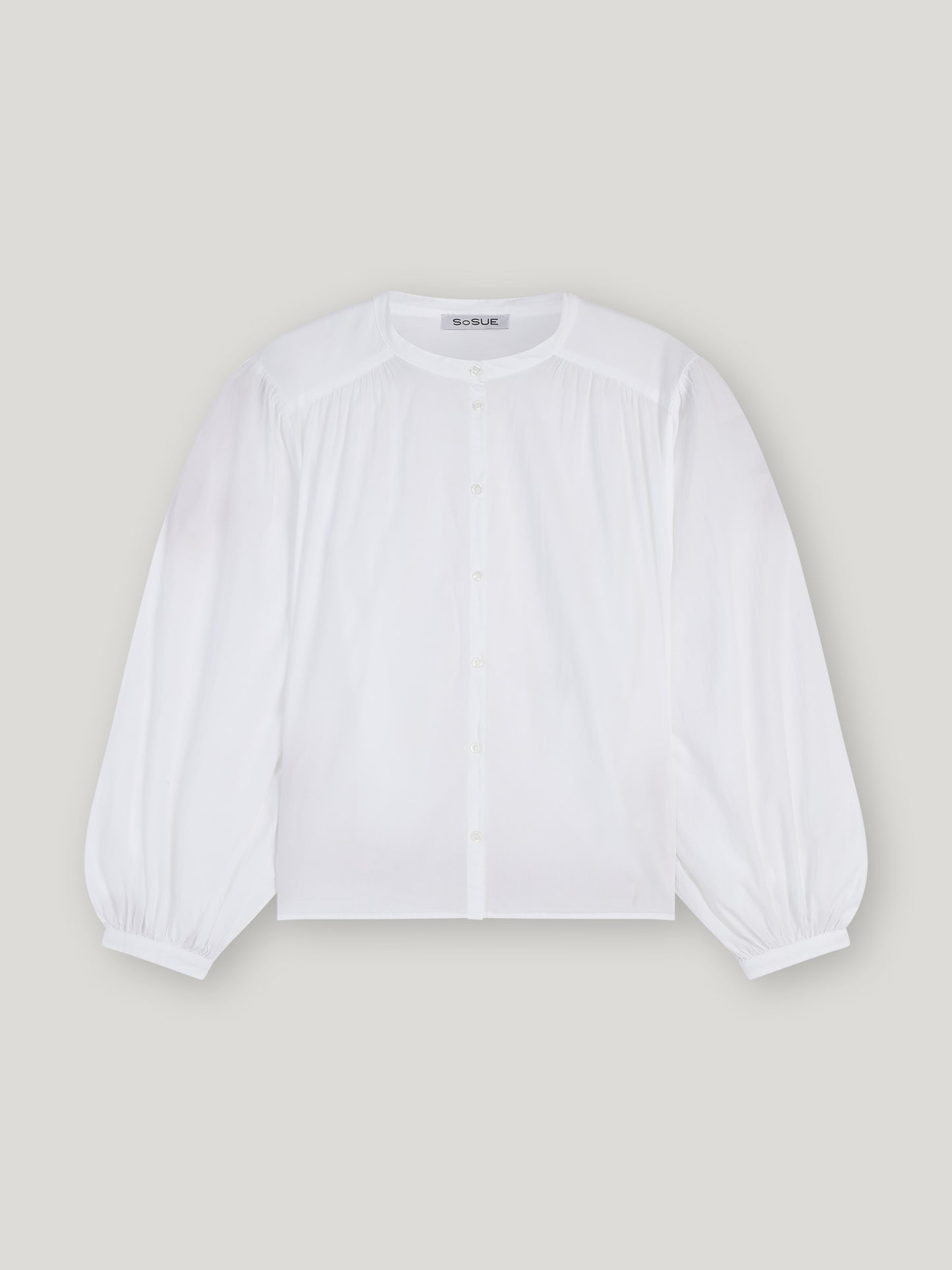 Die Bluse Bella White hat einen Rundhalsausschnitt, eine geknöpfte Vorderseite, geraffte Schultern und Puffärmel mit geknöpften Manschetten in einer übergroßen Silhouette, die sich vor einem hellgrauen Hintergrund präsentiert.