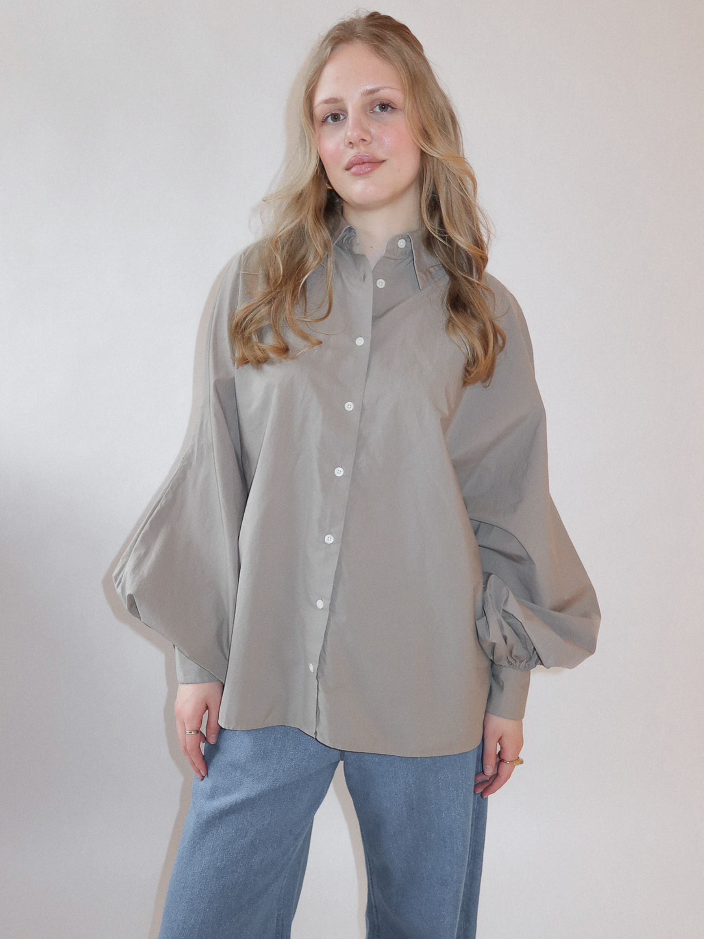 Eine Frau mit langen blonden Haaren trägt die übergroße Bluse Antonia Mud in hellgrau-beige mit Fledermausärmeln und hellblaue Jeans vor einem schlichten hellen Hintergrund.
