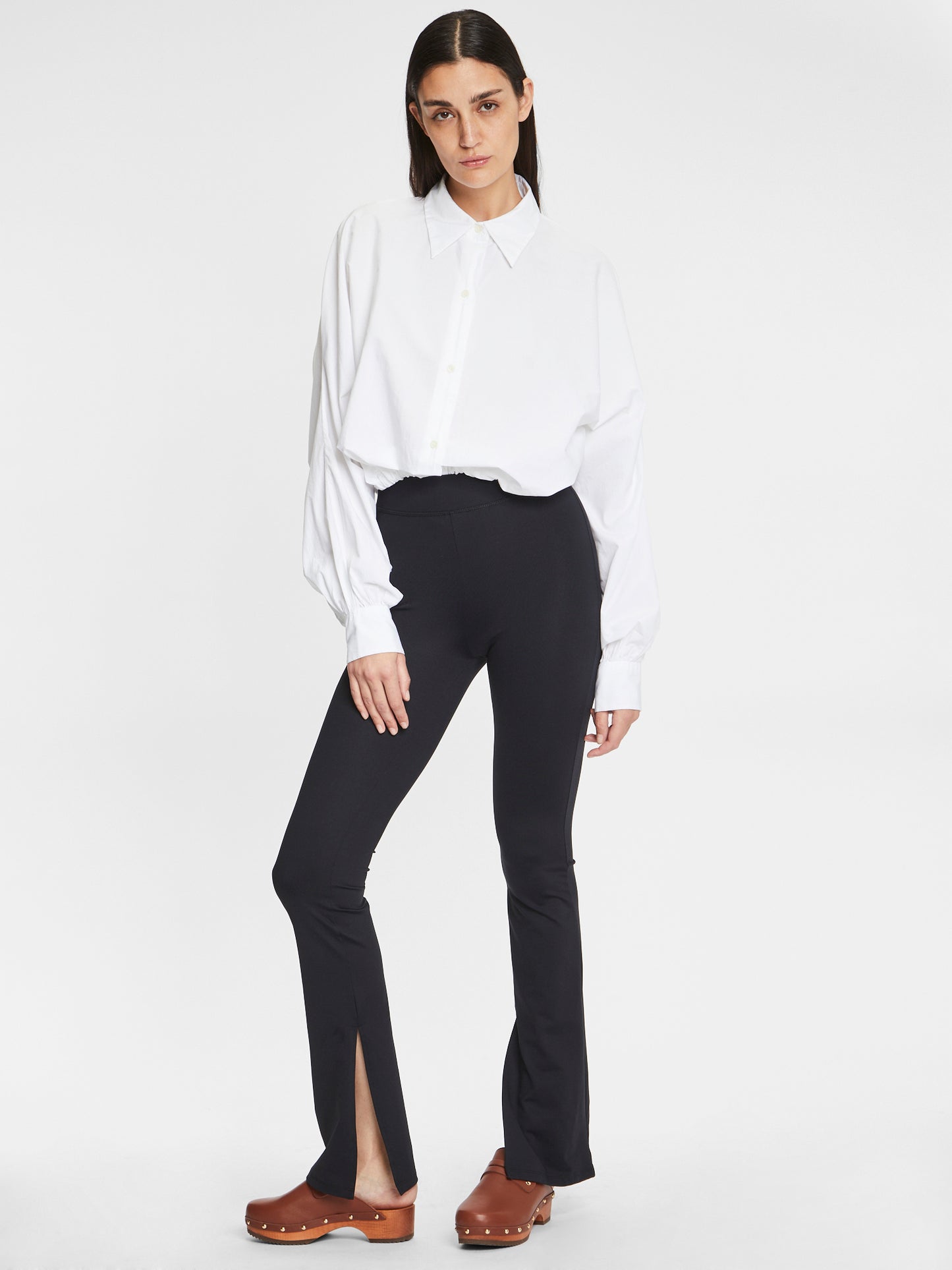 Eine Person mit langen dunklen Haaren steht vor einem weißen Hintergrund in einem lockeren weißen Hemd und Hose Slit Leggings-80% Recycled, mit Schlitzen vorne am Saum, gepaart mit braunen Clogs. Sie hat einen neutralen Gesichtsausdruck.