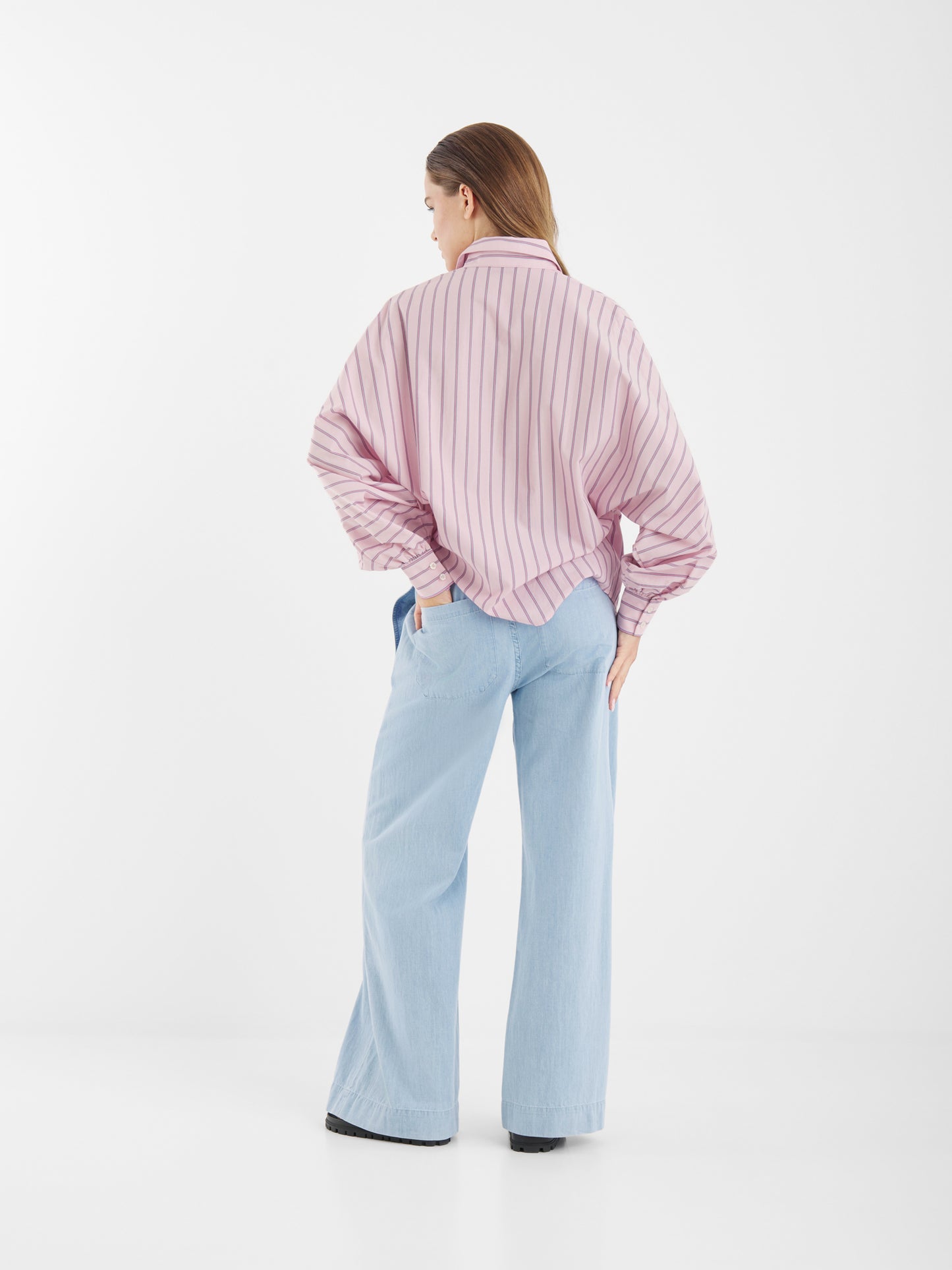 Eine Frau steht mit dem Antonia Slim Stripes Rose Hemd - locker, rosa-weiß gestreift mit weiten Ärmeln - und einer hellblauen Jeans mit weitem Bein. Eine Hand steckt in ihrer Hosentasche vor einem schlichten weißen Hintergrund.