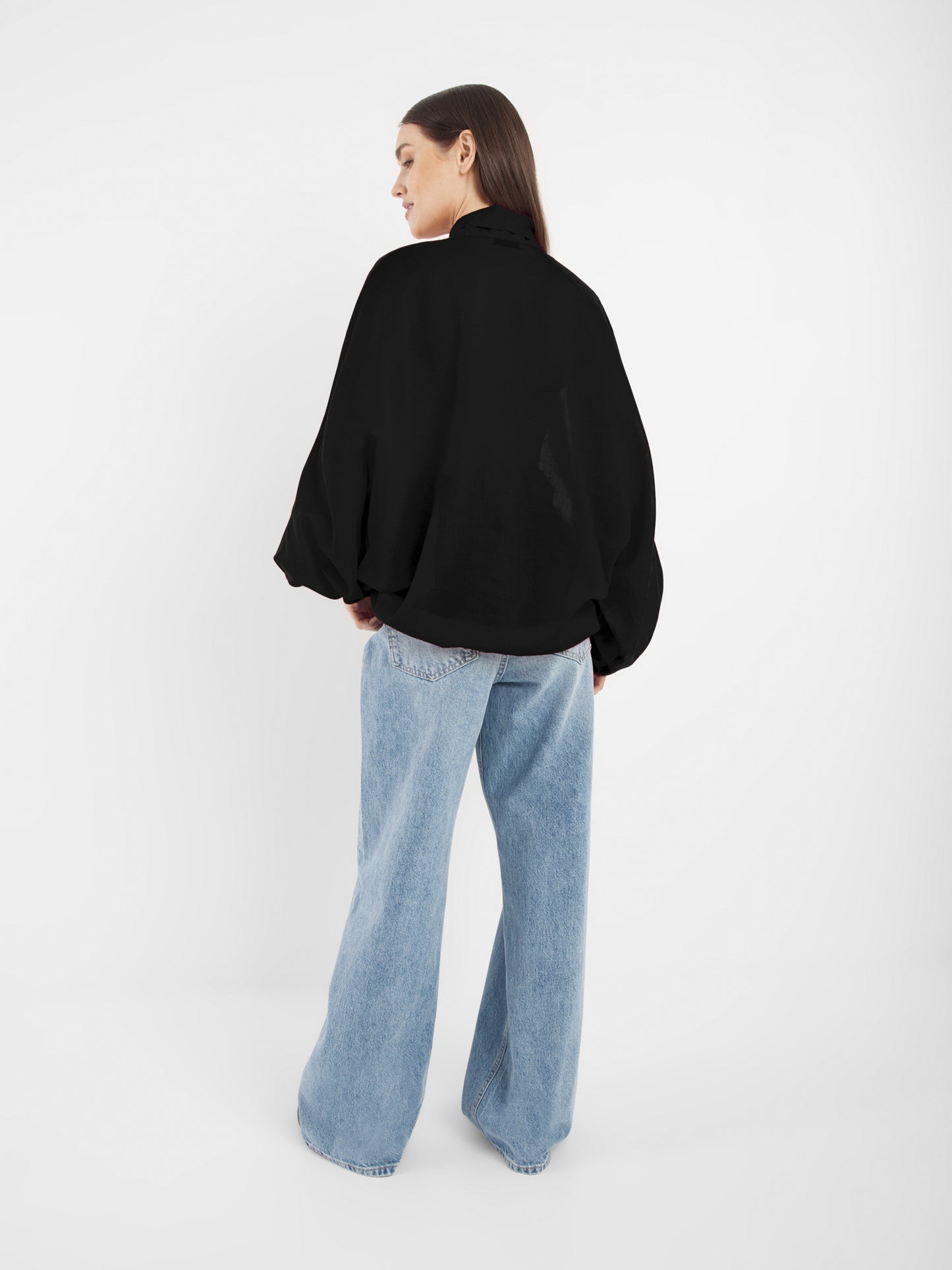 Eine Person, die sich abwendet, trägt die Bluse Antonia Bow Taupe mit langen Ärmeln und hellblaue Jeans mit weitem Bein vor einem schlichten weißen Hintergrund.