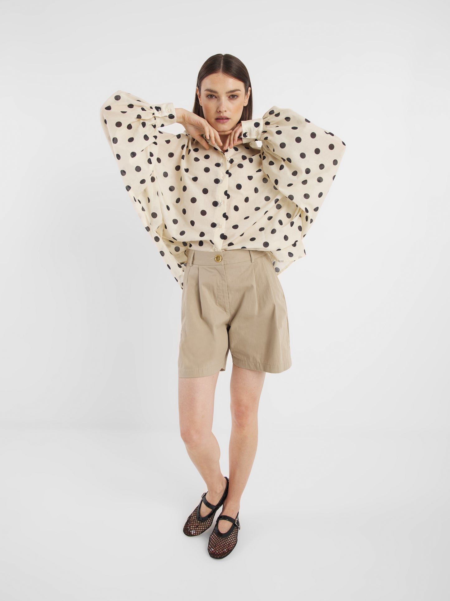 Eine Frau posiert vor einem schlichten weißen Hintergrund und trägt die Bluse Antonia Dots Ecru Black, eine übergroße Baumwoll-Voile-Bluse mit schwarzen Tupfen, gepaart mit beigen Shorts mit hoher Taille und schwarzen Netzschuhen. Ihre Arme sind in der Nähe ihres Kinns erhoben, während sie in die Kamera schaut.