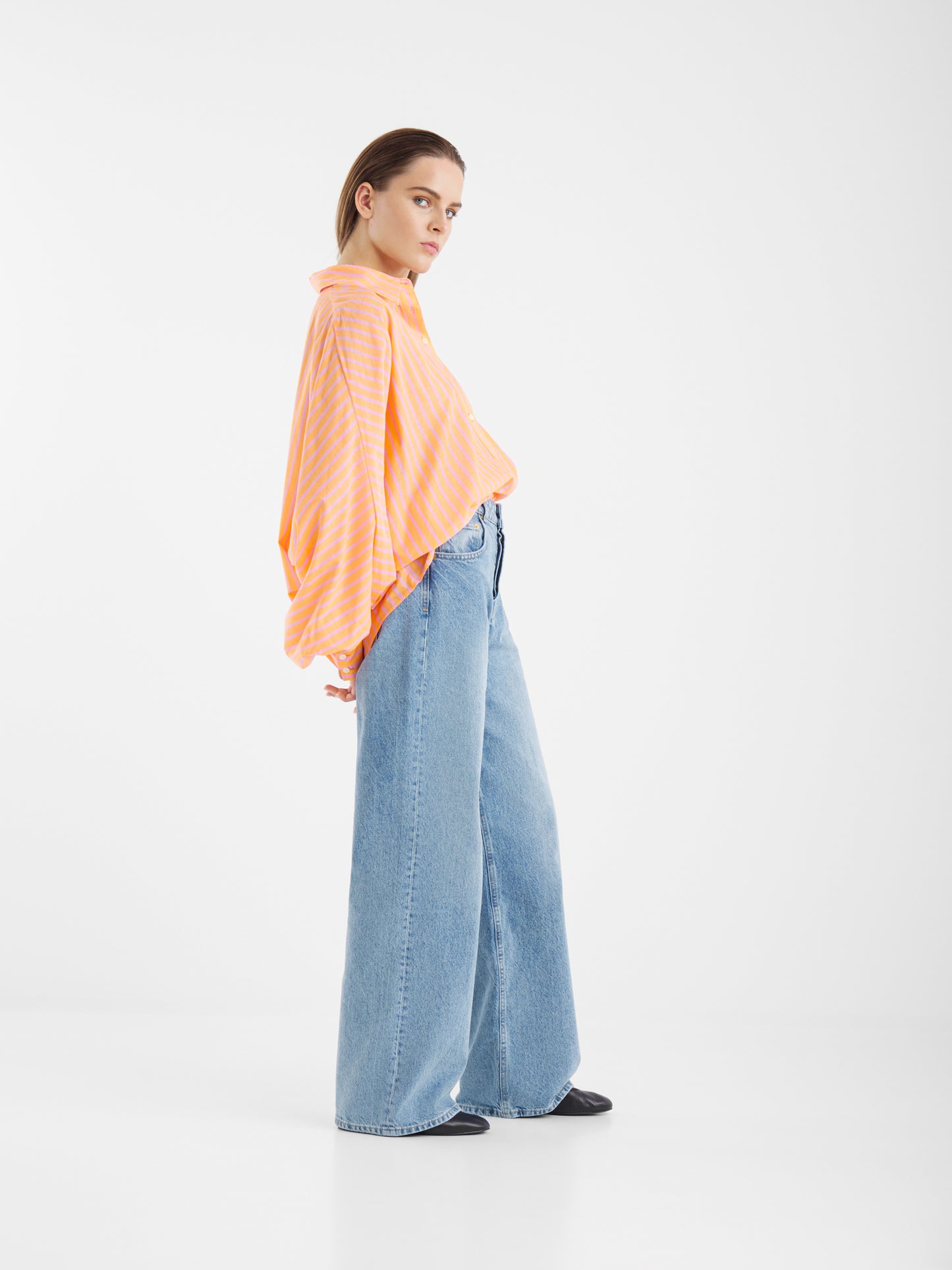 Eine Person steht seitlich vor einem schlichten weißen Hintergrund und trägt das Hemd Antonia Voile Coral Pink, das locker mit einer weit geschnittenen Bluejeans und schwarzen Schuhen kombiniert ist. Ihre Hände sind hinter dem Rücken verschränkt und sie hat einen neutralen Gesichtsausdruck.