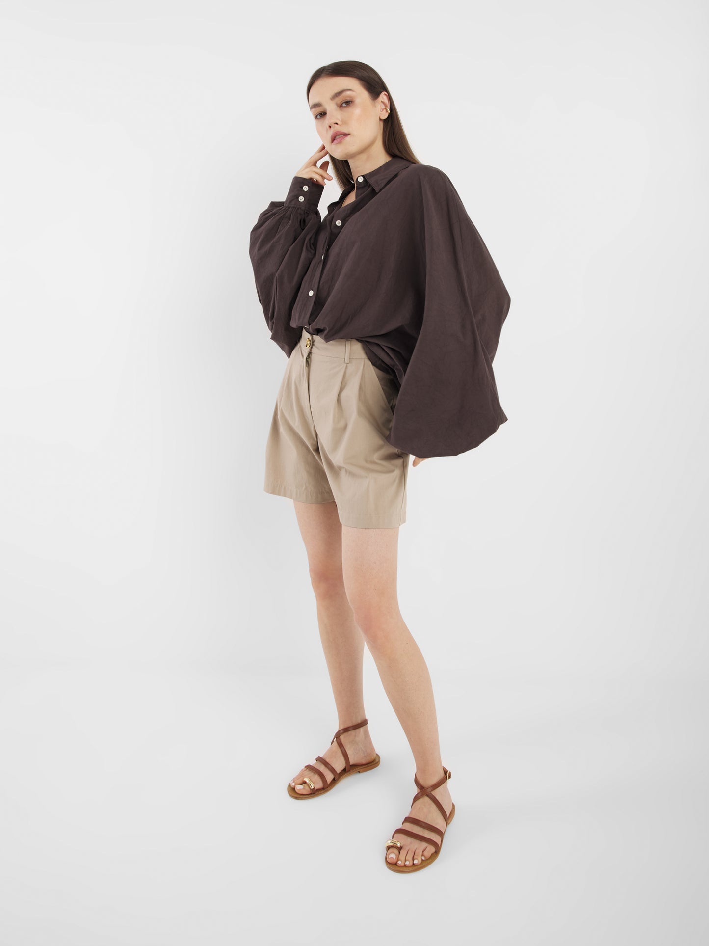 Eine Frau posiert vor einem weißen Hintergrund und trägt das lockere Hemd Antonia Cotton Brown mit Knopfleiste, beigefarbene Shorts mit hoher Taille und braune Riemchensandalen. Sie steht mit einer Hand neben ihrem Gesicht und sieht entspannt aus.