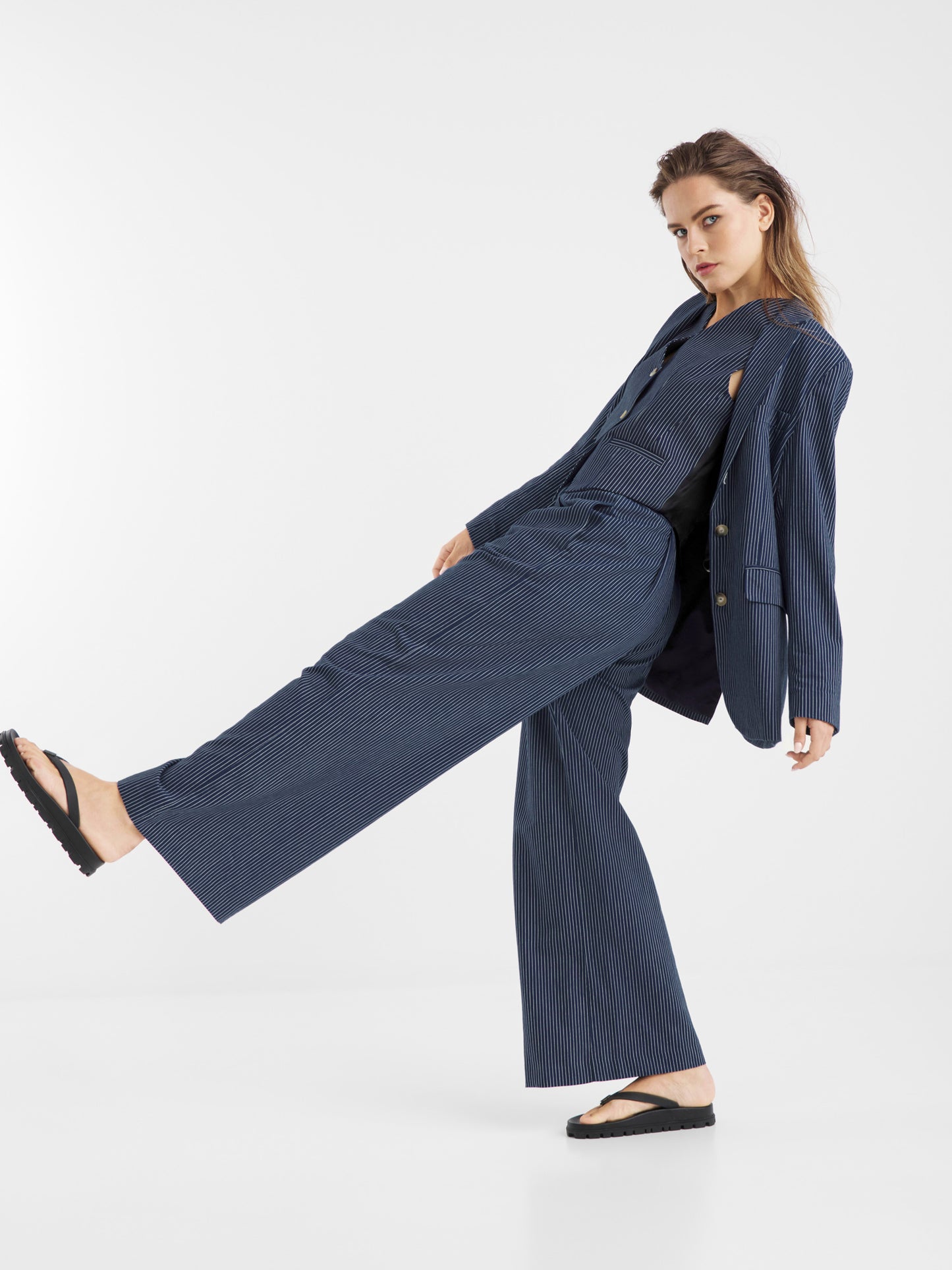 Eine Frau, die einen locker sitzenden Blazer mit Nadelstreifen in Navy und schwarze Sandalen trägt, posiert mit einem angehobenen und angewinkelten Bein vor einem schlichten weißen Hintergrund.