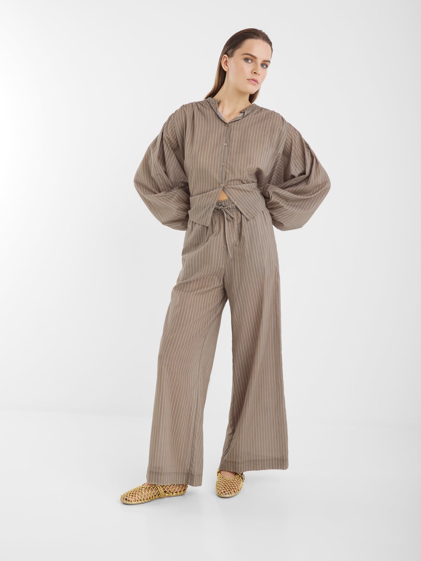 Eine Frau steht in einem neutralen Atelier und trägt eine taupefarbene Nadelstreifenhose mit weitem Bein und die luftige Bluse Bella Taupe Stripes mit weiten Ärmeln, gepaart mit gewebten gelben Schuhen.