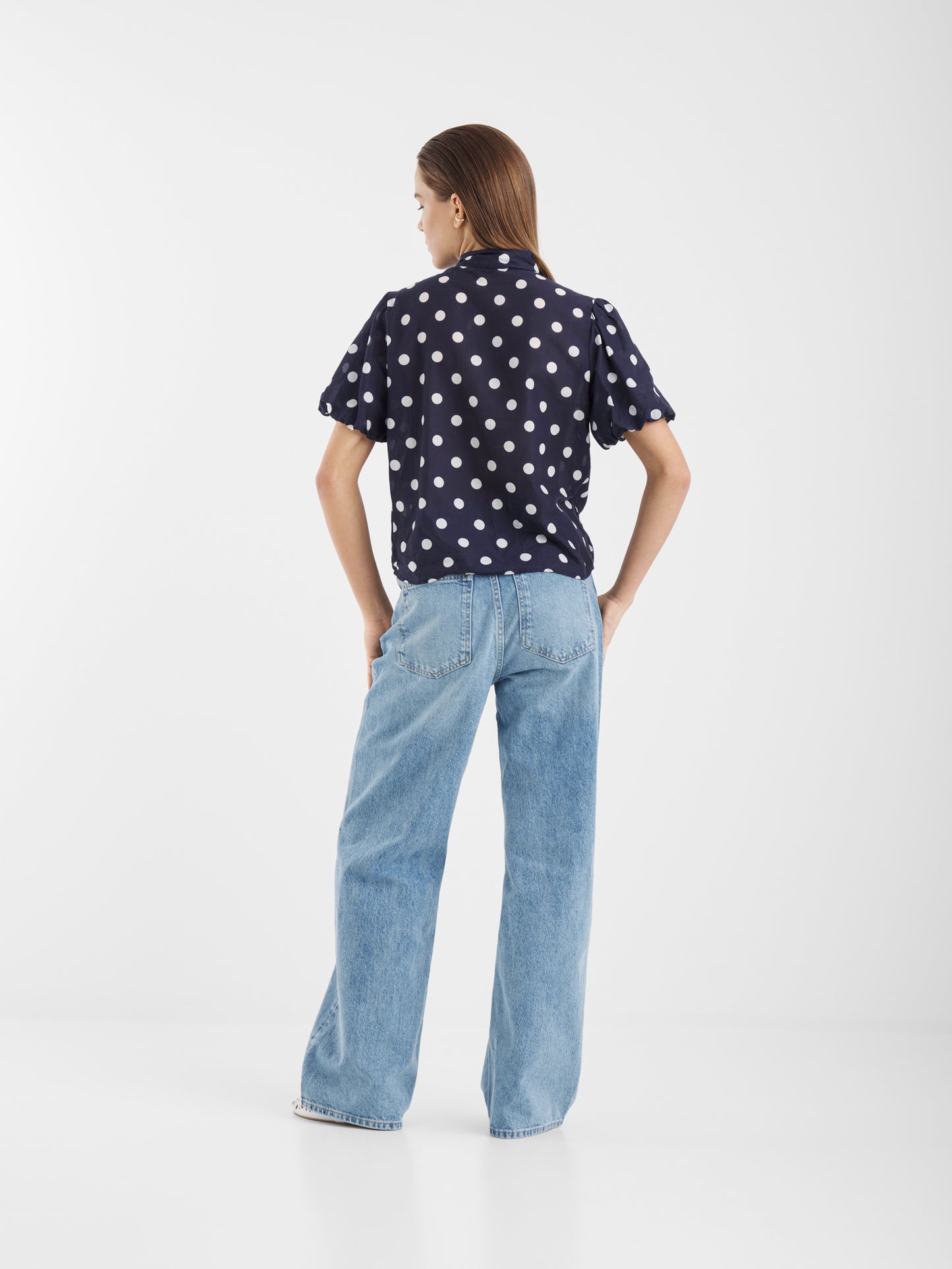 Eine Frau steht mit der Bluse Bow Slim Dots Navy Off White und hellblauen, weit geschnittenen Jeans vor einem schlichten weißen Hintergrund.