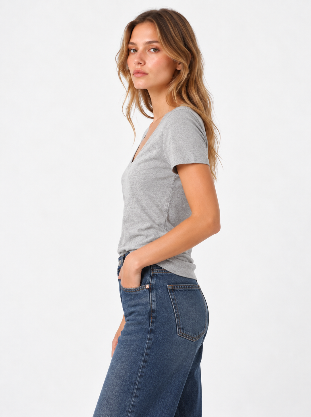 Eine Frau mit langen, gewellten Haaren steht im Profil vor einem weißen Hintergrund. Sie trägt ein graues T-Shirt mit V-Ausschnitt und eine blaue Jeans, hat die Hände in den Taschen und blickt selbstbewusst in die Kamera.
