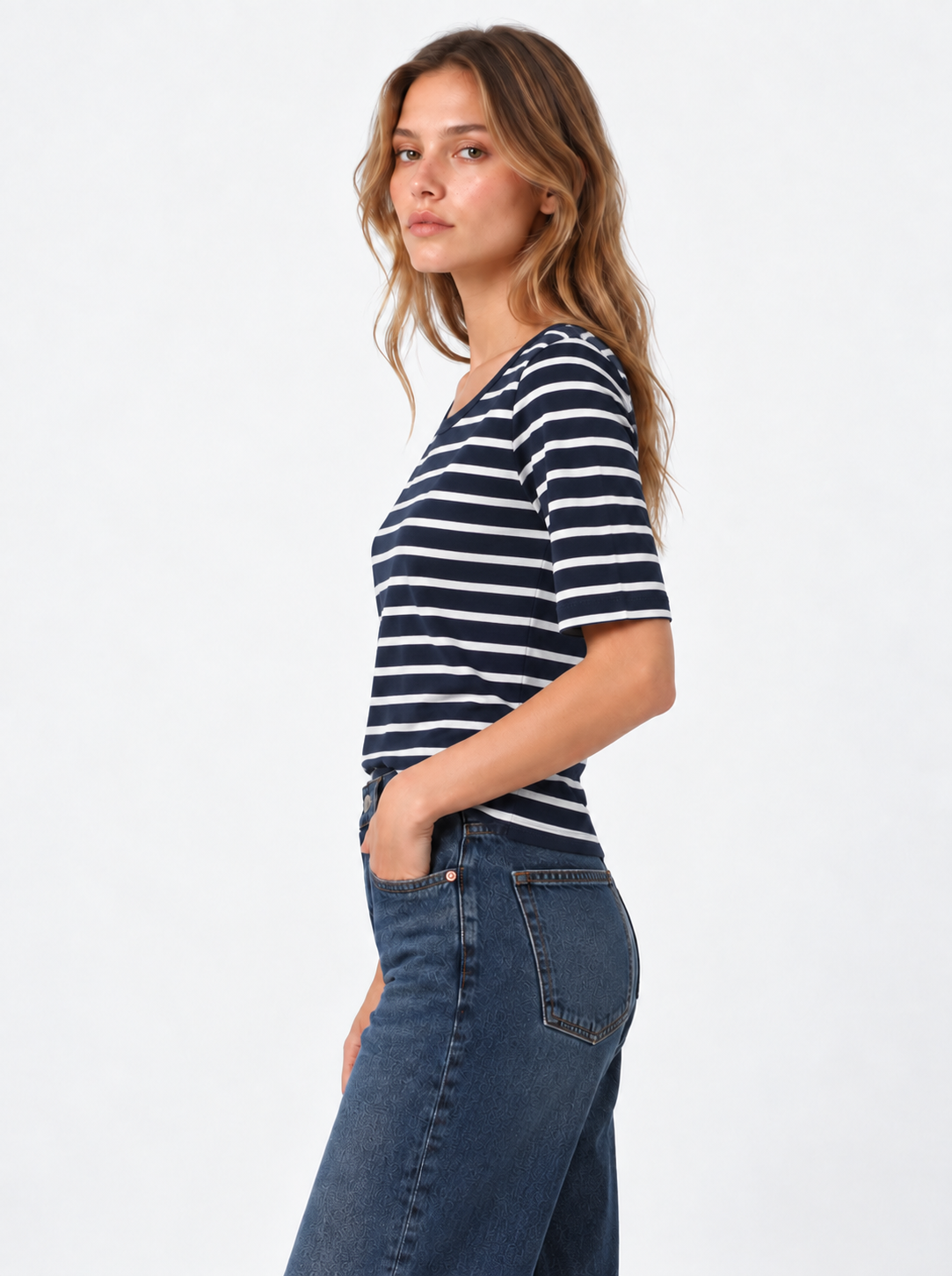 Eine Frau, die ein T-Shirt in Marineblau mit weißen Streifen im klassischen Marinière-Stil und eine blaue Jeans trägt, steht im Profil mit der linken Hand in der Tasche vor einem schlichten, hellen Hintergrund.