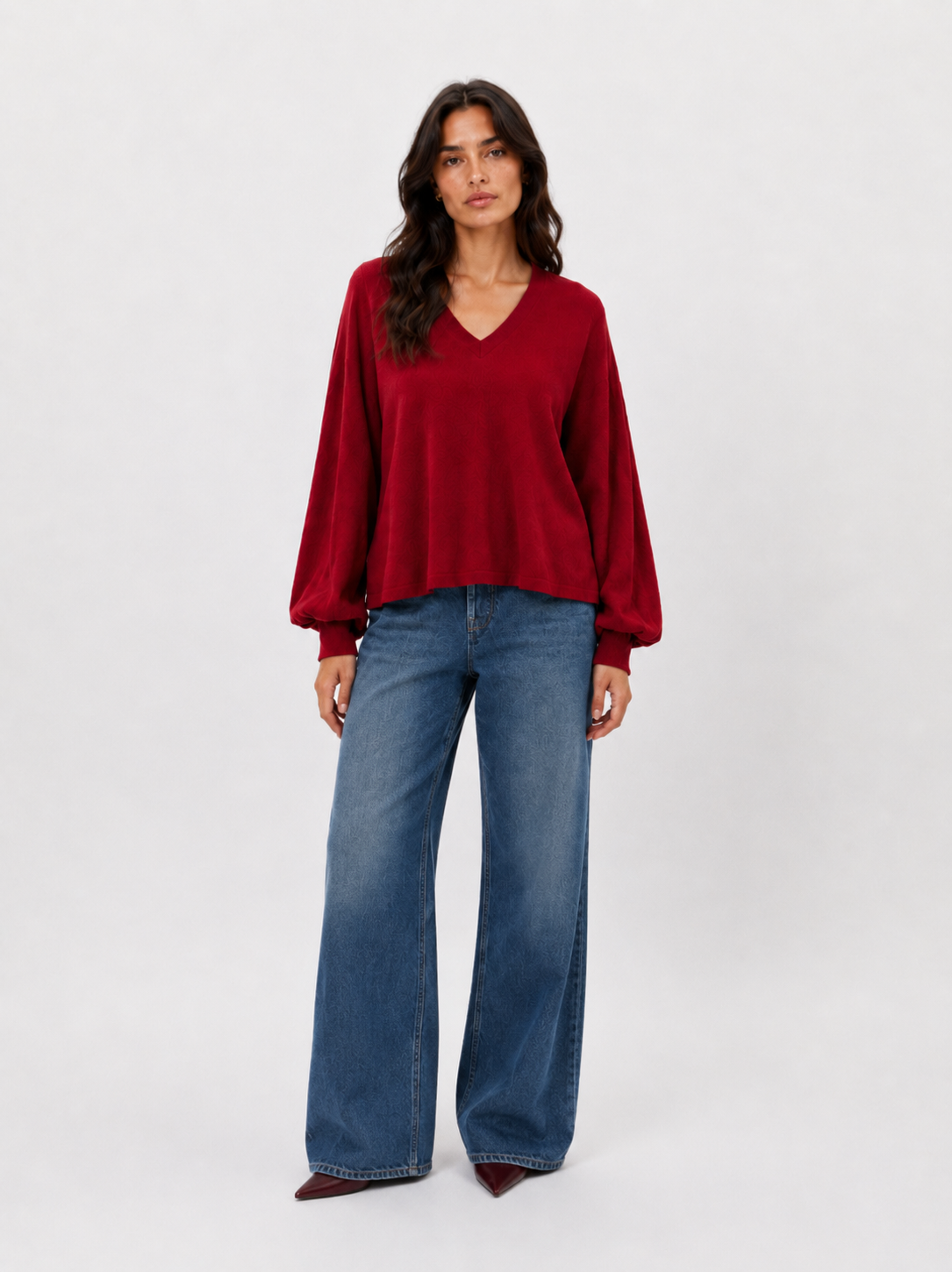 Eine Frau steht mit dem Blick nach vorne und trägt einen locker sitzenden, langärmeligen Pullover „Antonia V-Neck Rot“ aus luxuriöser Merinowolle und Kaschmir, dazu eine blaue Jeans mit weitem Bein und Schuhe mit spitzer Zehenpartie. Der Hintergrund ist einfarbig und hell.