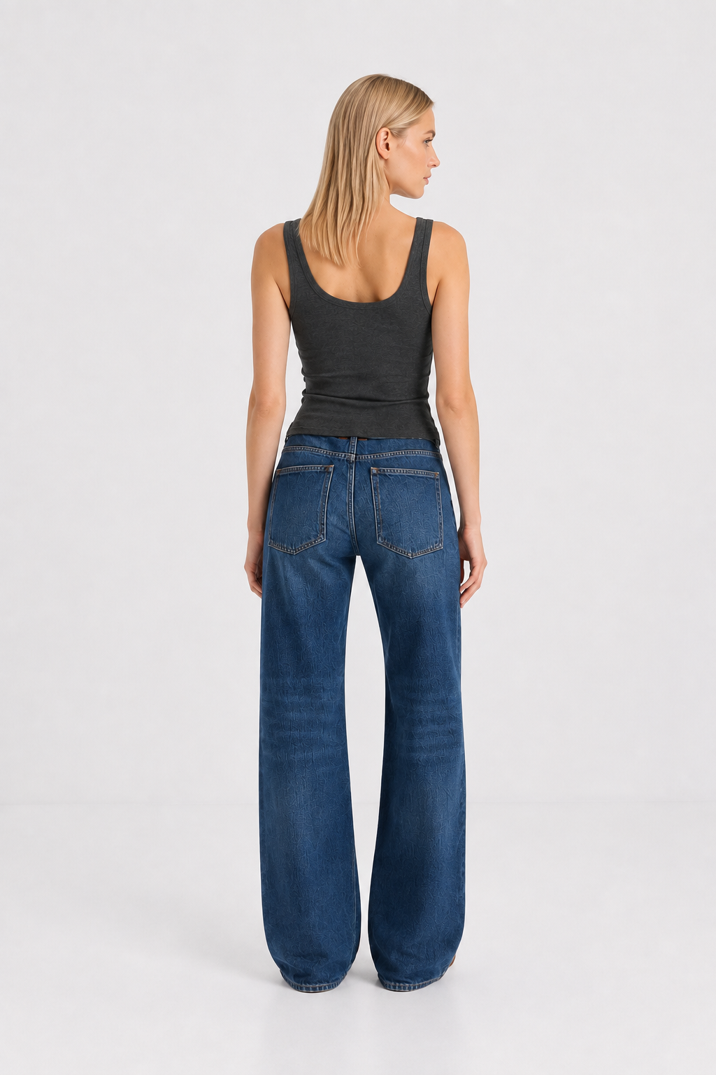 Eine Frau steht mit dem Rücken zur Kamera und trägt das figurbetonte Tanktop „Anthrazit“ sowie eine hoch taillierte Bluejeans mit weitem Bein. Sie hat glattes blondes Haar und posiert vor einem schlichten weißen Hintergrund.