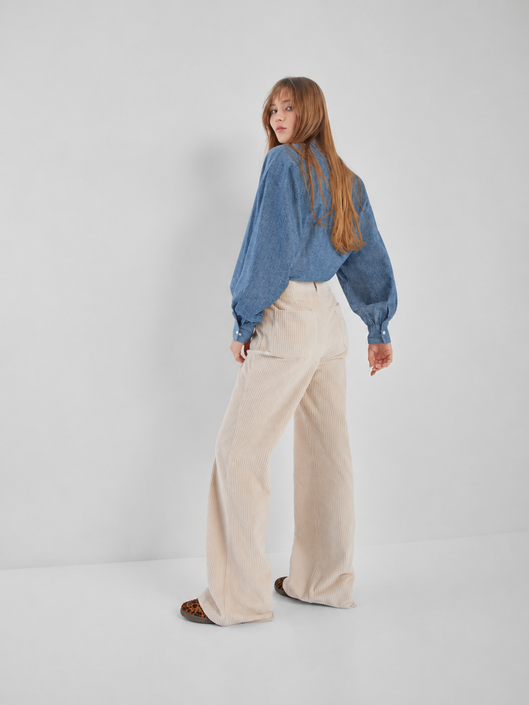 Eine Frau mit langen Haaren trägt die Bluse „Antonia Slim Denim“ und eine cremefarbene Cordhose mit hoher Taille. Sie steht in einer minimalistischen Kulisse und blickt über ihre Schulter.