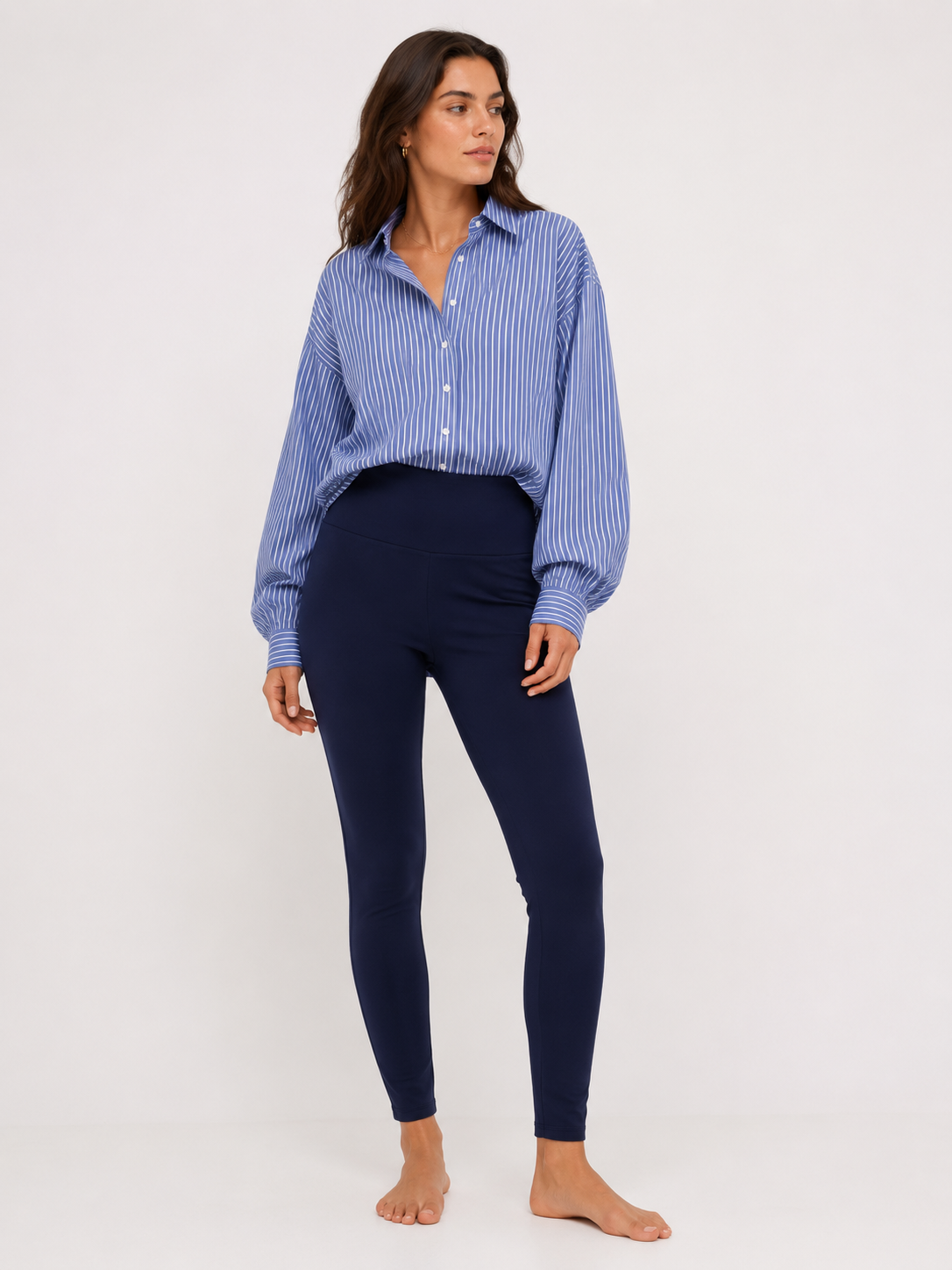 Eine Frau steht barfuß vor einem schlichten Hintergrund und trägt ein blau-weiß gestreiftes Hemd mit Knöpfen, weiten Ärmeln und eine hoch taillierte Leggings in Marineblau. Sie blickt mit ausdruckslosem Gesicht zur Seite, wodurch die Dehnbarkeit ihrer Leggings besonders zur Geltung kommt.