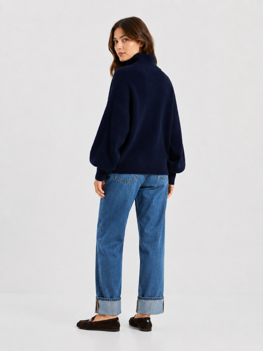 Eine Frau steht mit leicht abgewandtem Blick da und trägt eine dunkelblaue Bomber-Strickjacke, eine blaue Jeans mit Umschlag und schwarze Schuhe. Vor einem schlichten, hellen Hintergrund blickt sie über ihre Schulter.