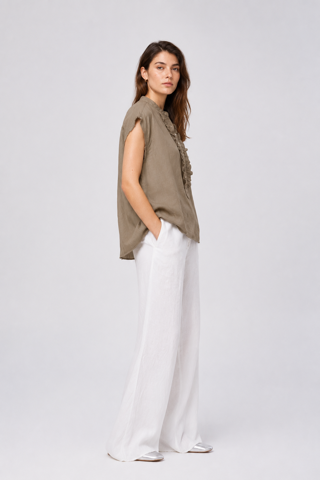 Eine Frau steht in einer entspannten Haltung vor einem schlichten, hellen Hintergrund und trägt eine locker sitzende Bluse „Barcelona Linen Soft Brown“ sowie eine weiße Hose mit weitem Bein und weiße Schuhe.