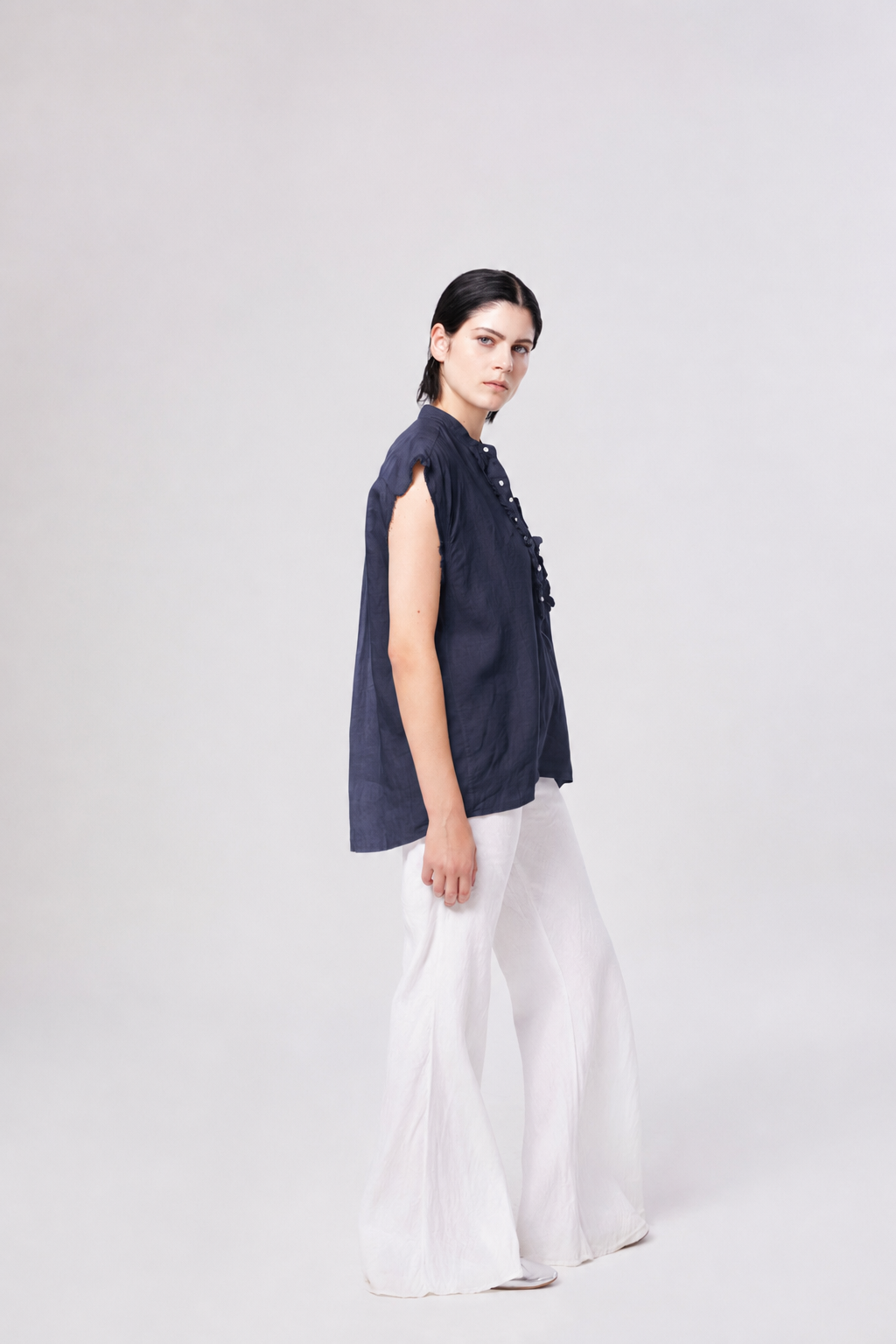 Eine Person mit kurzen, dunklen Haaren steht seitlich vor einem schlichten, hellen Hintergrund und trägt eine lockere, marineblaue Bluse von Barcelona Linen sowie eine weiße Hose mit weitem Bein.