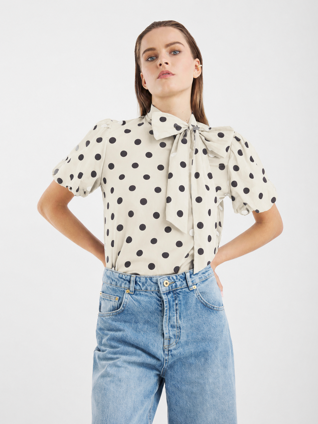 Eine Person trägt die Bluse „Bow Dots Ecru Black“ – eine cremefarbene Bluse aus Baumwollvoile mit schwarzen Punkten, kurzen Puffärmeln und einer großen Schleife am Hals – kombiniert mit hellblauen Jeans mit hoher Taille, und steht vor einem schlichten weißen Hintergrund.